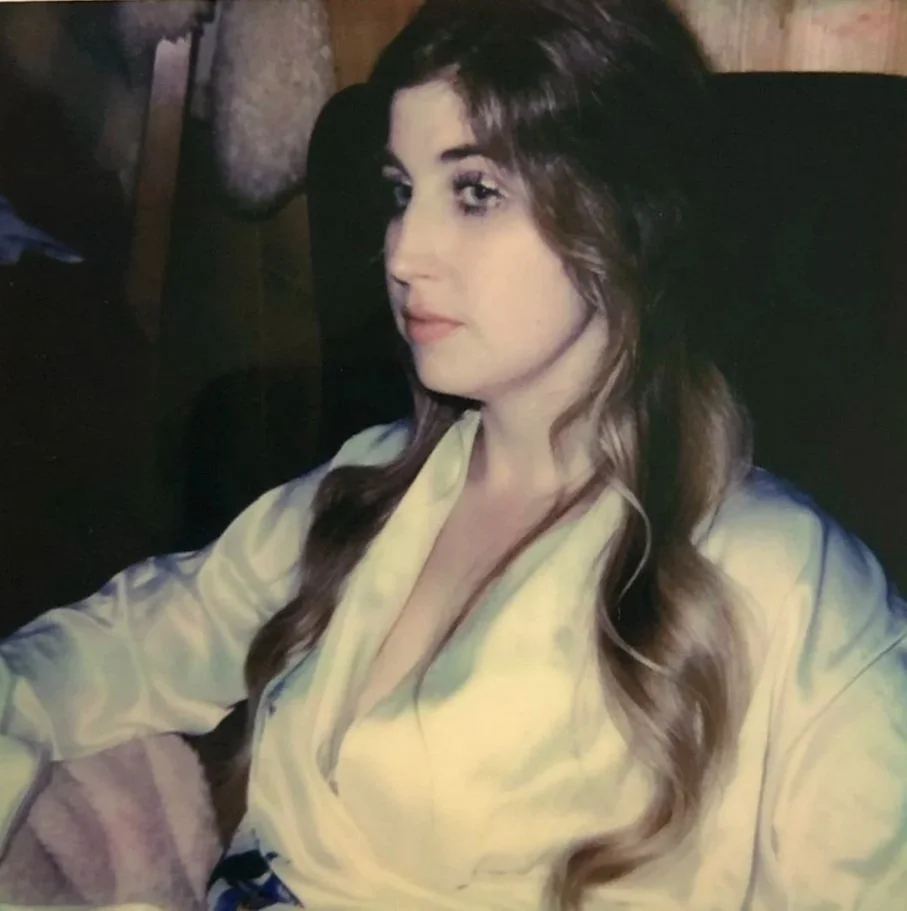 A woman with long, wavy brown hair, wearing a yellow satin blouse, sits in a dimly lit room with wooden walls.