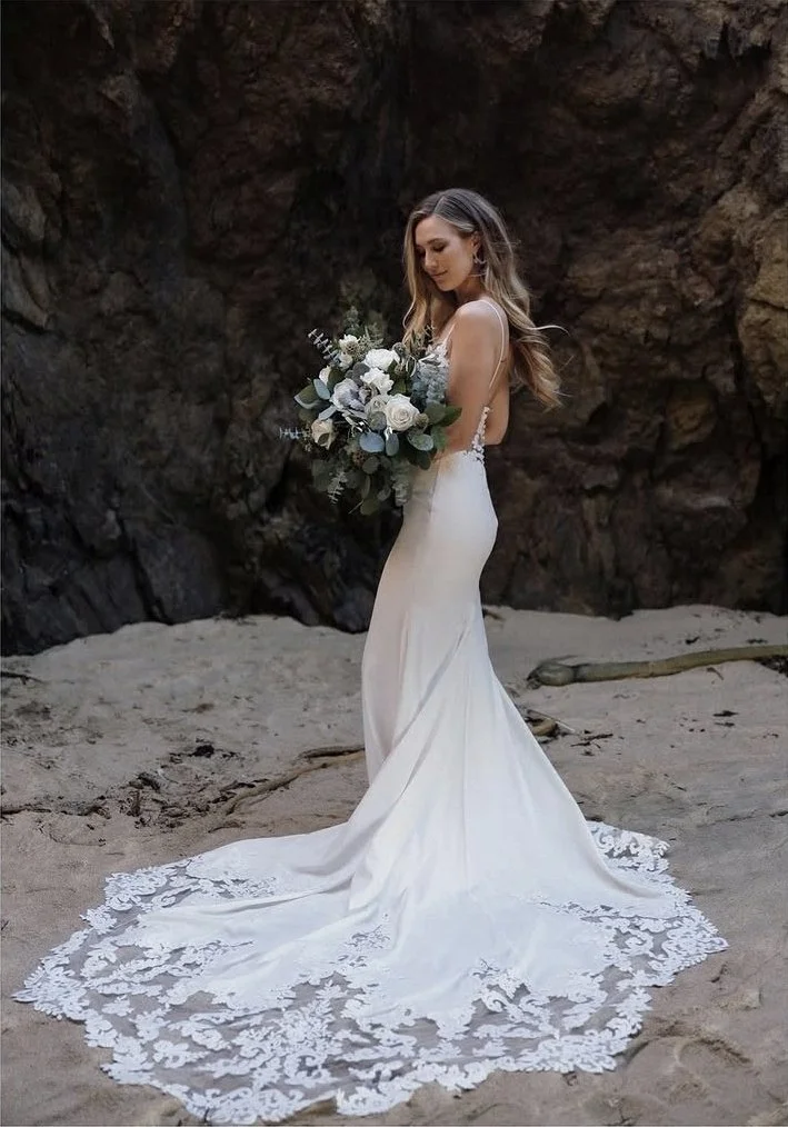A woman in a white wedding dress with a lace train holding a large bouquet of white and green flowers, standing on a sandy beach with rocky cliffs in the background.