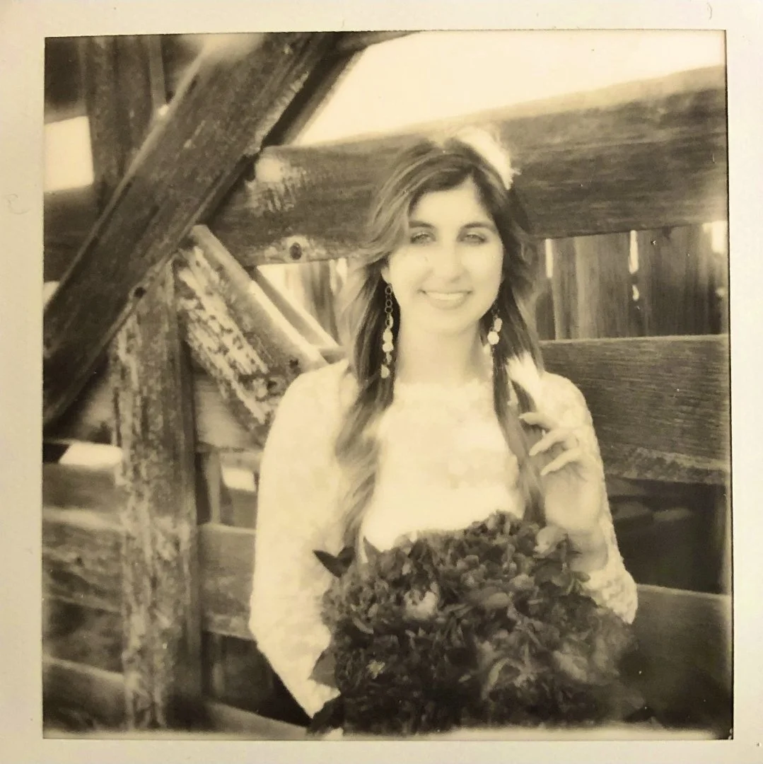 A woman with long wavy hair, wearing earrings and holding a bouquet, smiling while sitting outdoors near wooden structures.