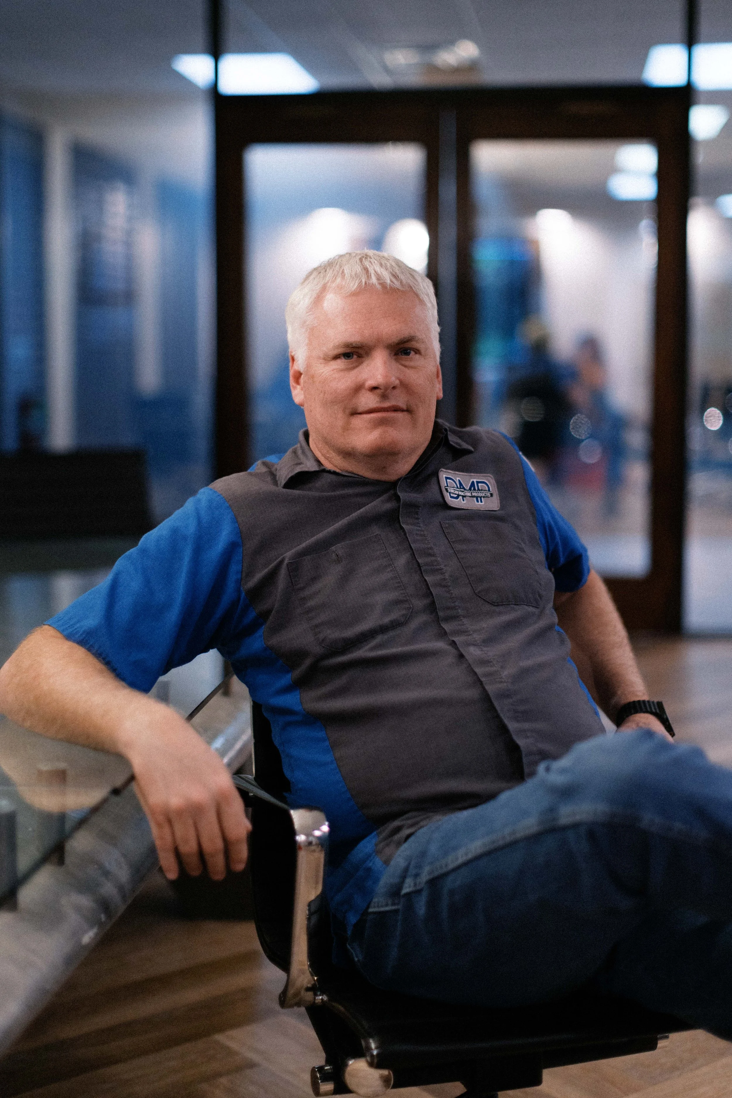 A man with short blonde hair sitting in an office chair in front of a glass door, wearing a gray and blue uniform with a logo, and looking at the camera.