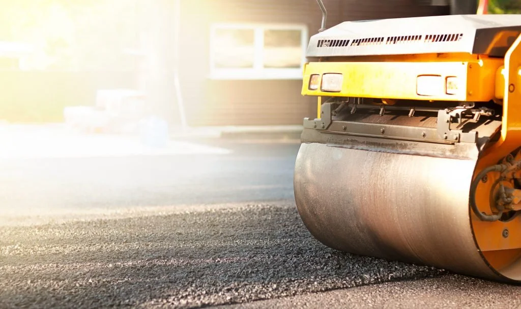 A yellow asphalt roller compacts a freshly paved road during daytime.