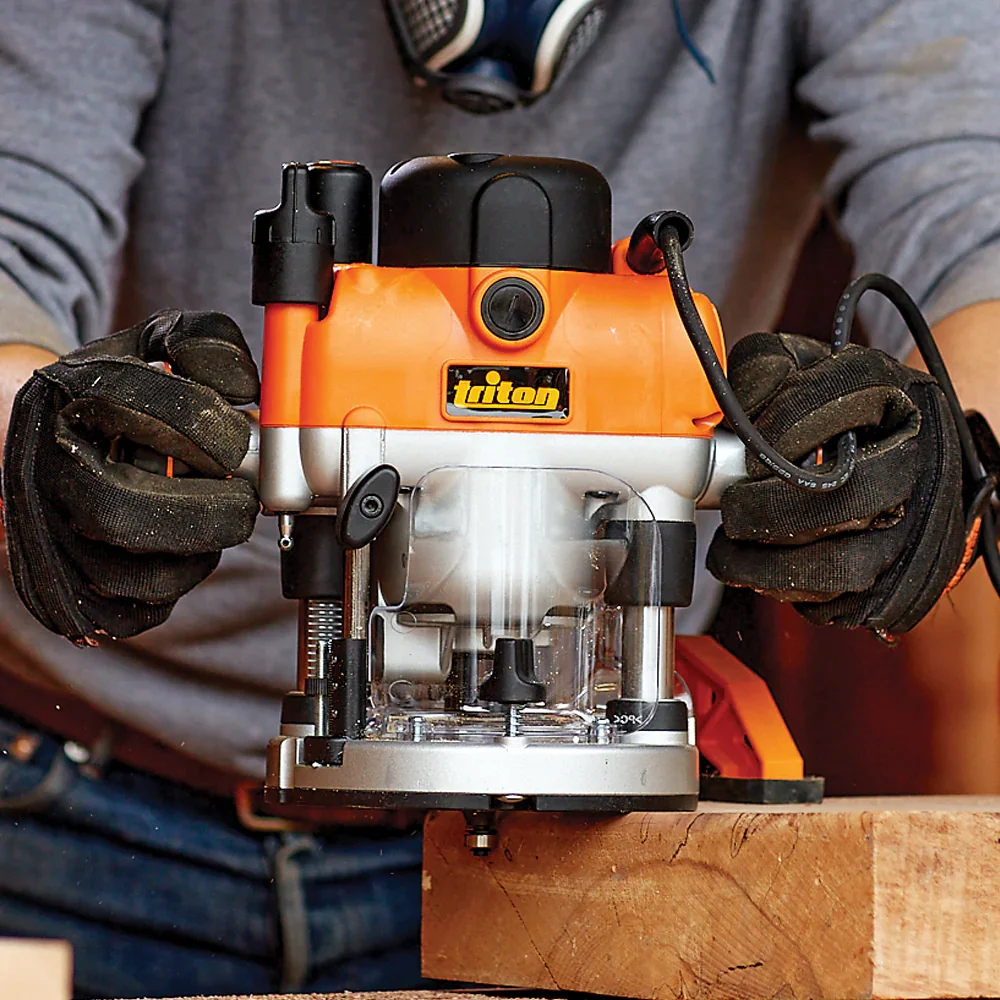 A person wearing gray sleeves and black gloves holding an orange and gray Triton router with a black power cord, operating on a piece of wood.