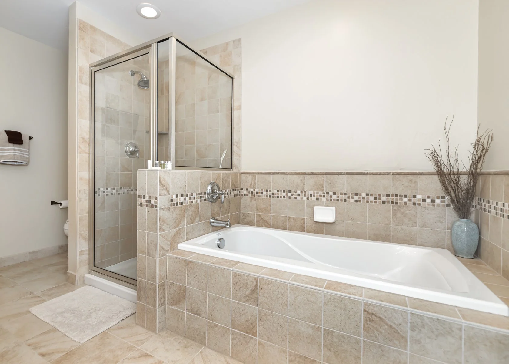 Bathroom with a glass-enclosed shower, a bathtub with beige tiles, a white soap holder, and a vase with dried branches in the corner.