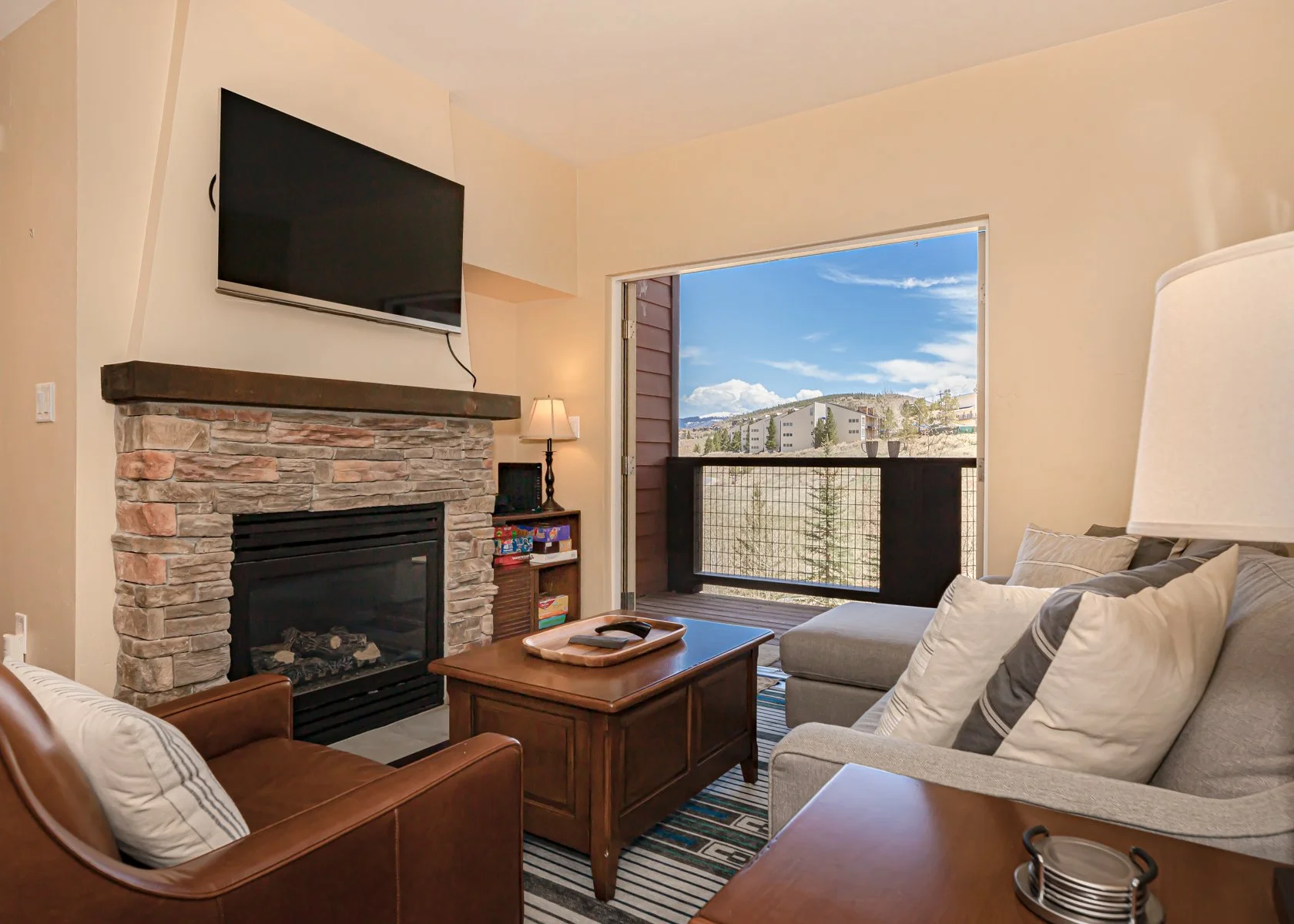 Living room with a stone fireplace, a wall-mounted TV, a sliding door leading to a balcony with a mountain view, and cushioned furniture with pillows.
