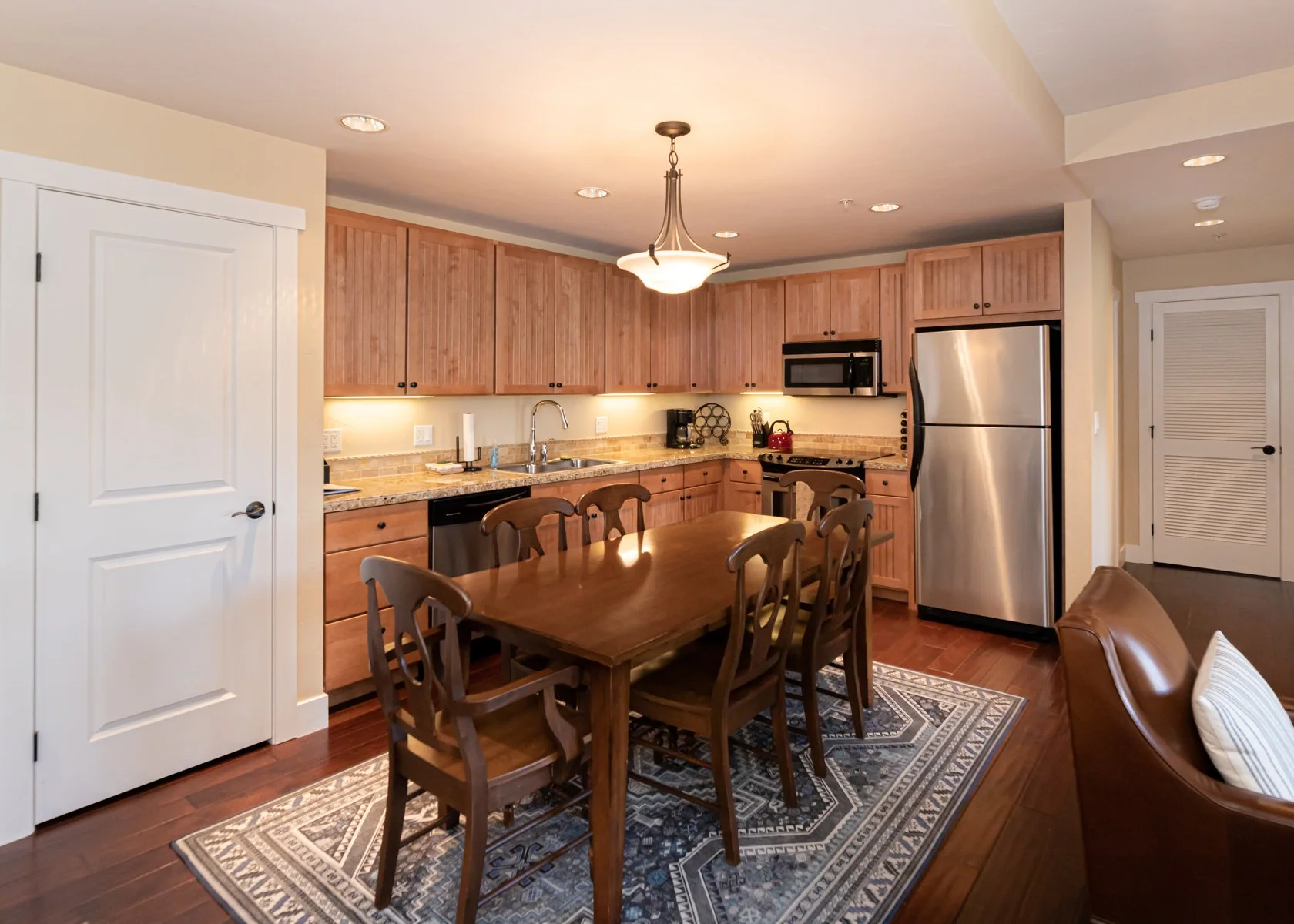 A kitchen with wooden cabinets, stainless steel refrigerator, microwave, and dishwasher. A wooden dining table with six chairs, area rug, and a ceiling light.