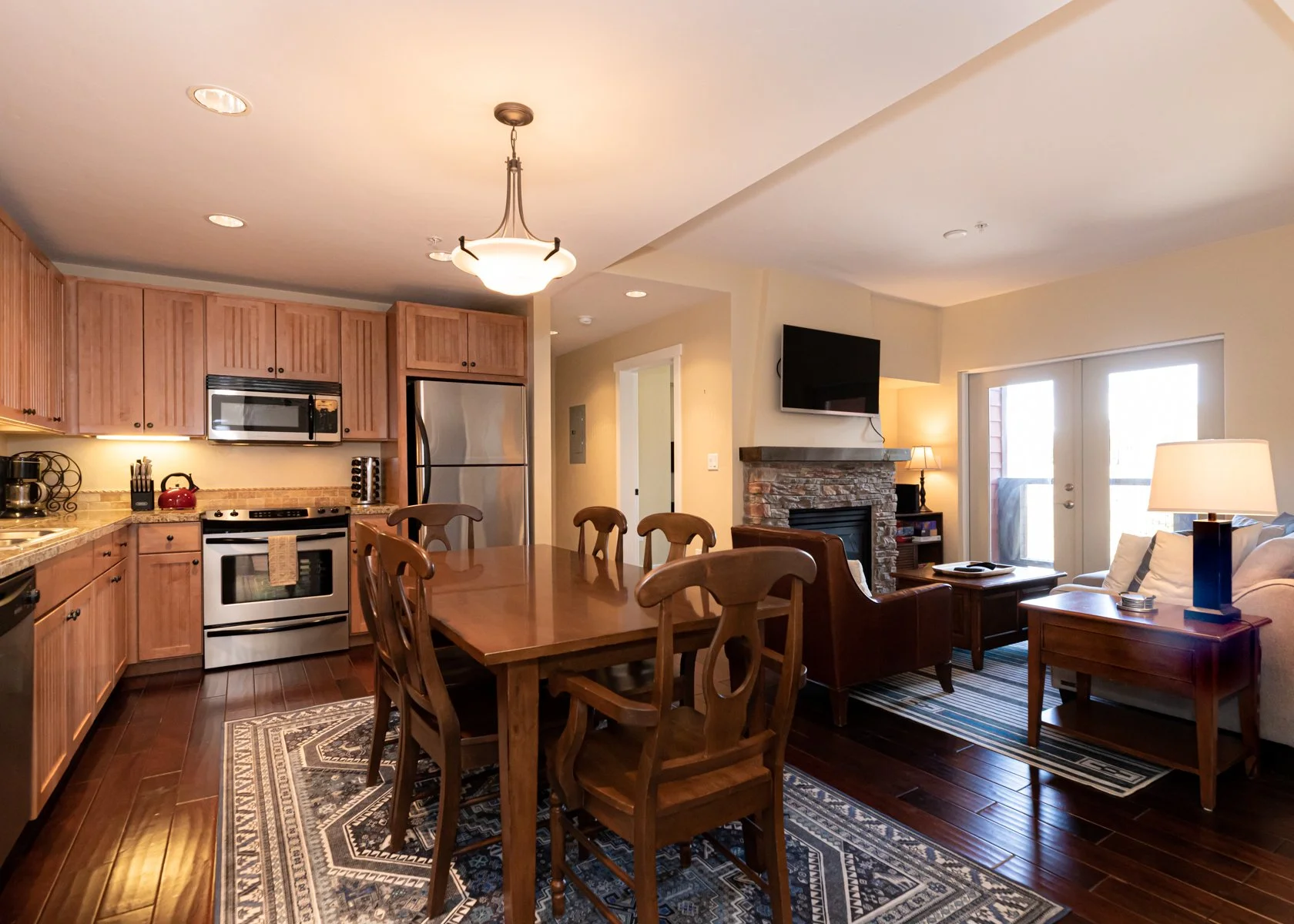 Open-concept living room and kitchen with wooden cabinetry, stainless steel appliances, a dining table with chairs, a stone fireplace, a sofa with a lamp, and sliding glass doors leading outside.