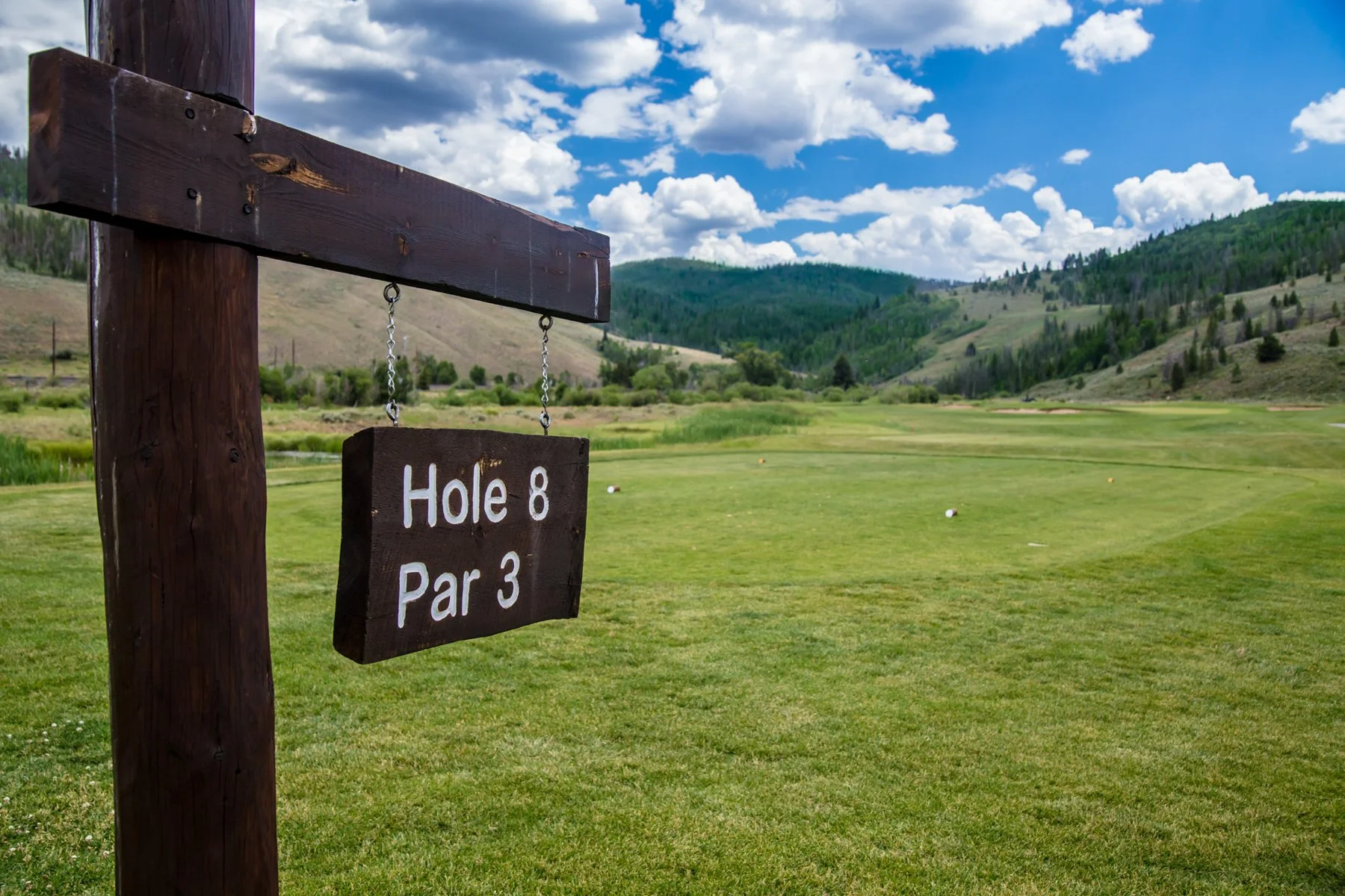 A wooden sign on a golf course indicating the eighth hole, a par 3, with lush green fairways and rolling hills in the background under a partly cloudy sky.