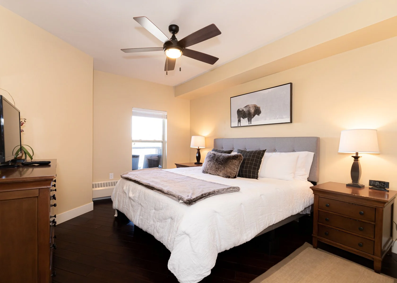 A cozy bedroom with a large bed, gray upholstered headboard, and white bedding. There are decorative pillows, a plush throw, and two nightstands with lamps. A framed bison picture hangs above the bed, with a window and a ceiling fan in the ceiling.
