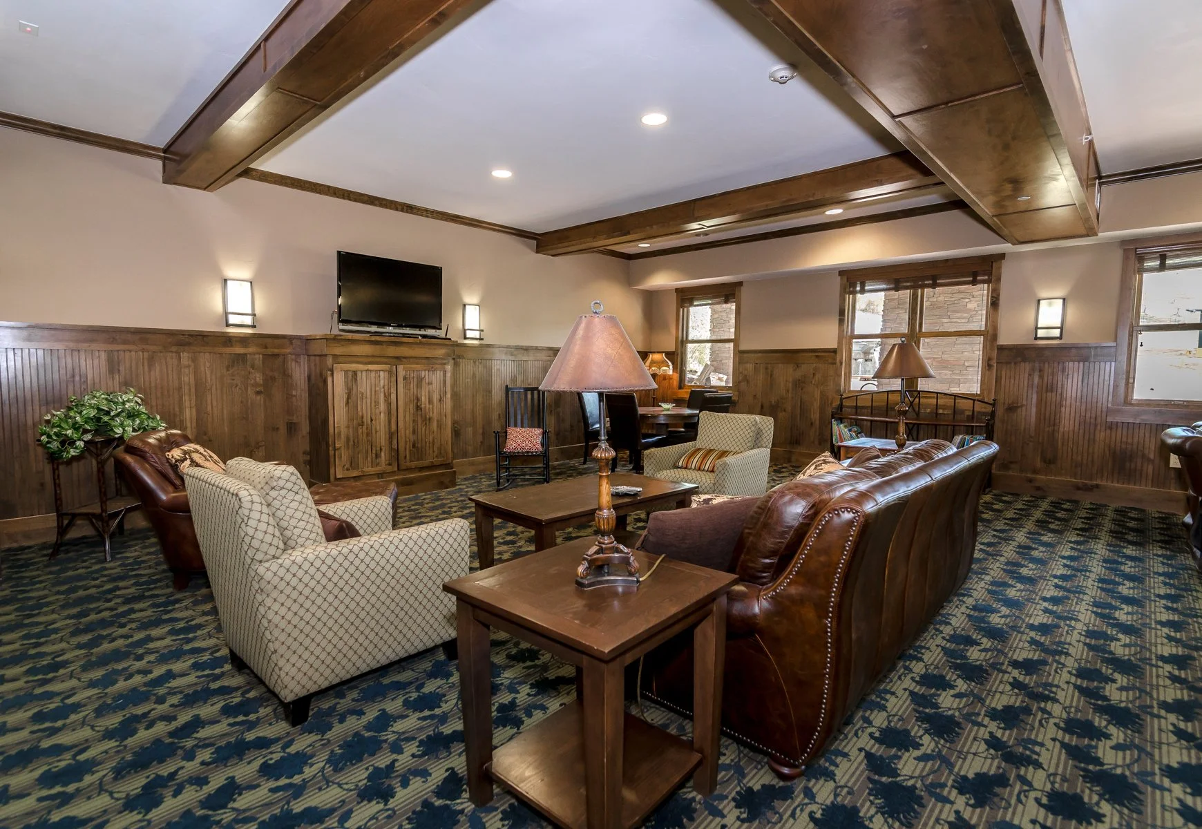 A cozy living room with wood-paneled walls and ceiling, featuring several cushioned chairs and a leather sofa arranged around a wooden coffee table, with lamps providing warm lighting and windows allowing natural light, on a patterned carpet.