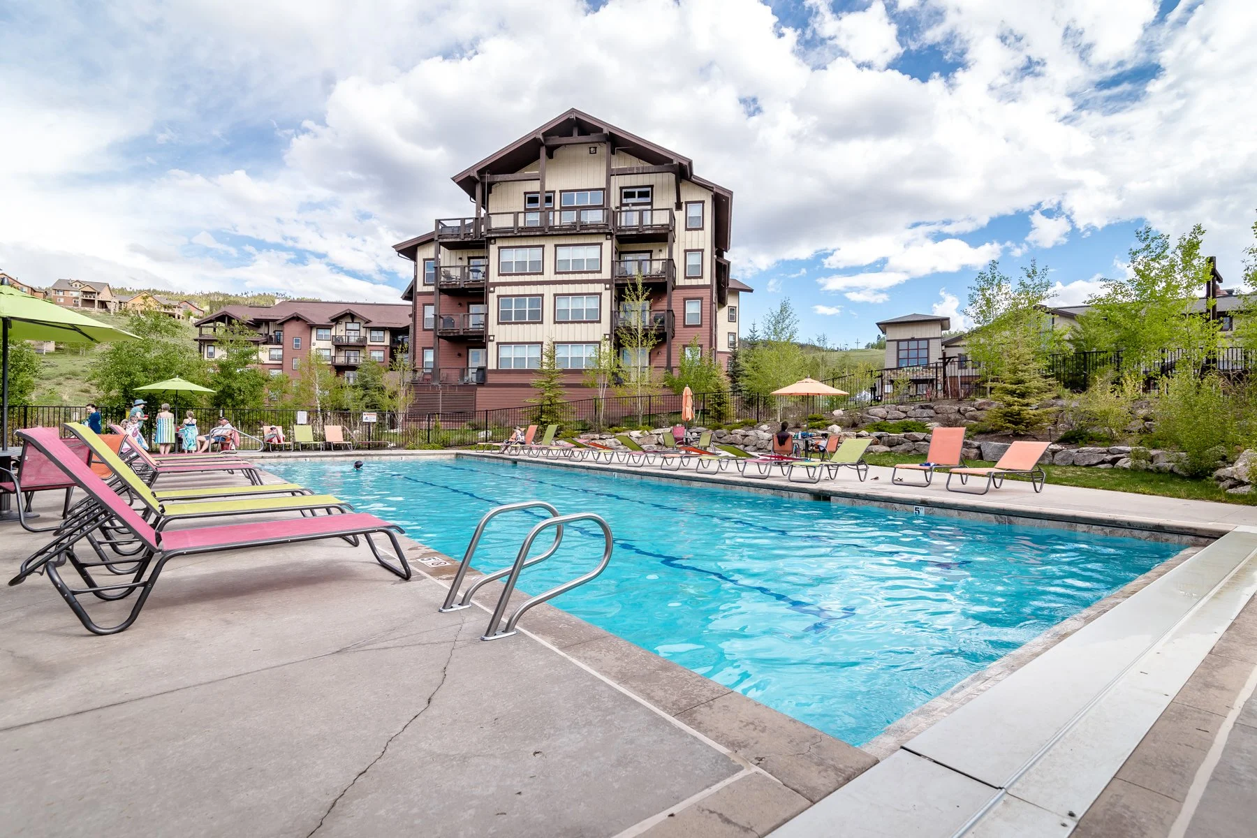 An outdoor swimming pool with a few people swimming and lounging on chairs. In the background, there is a multi-story residential building with balconies, surrounded by trees, with a partly cloudy sky overhead.
