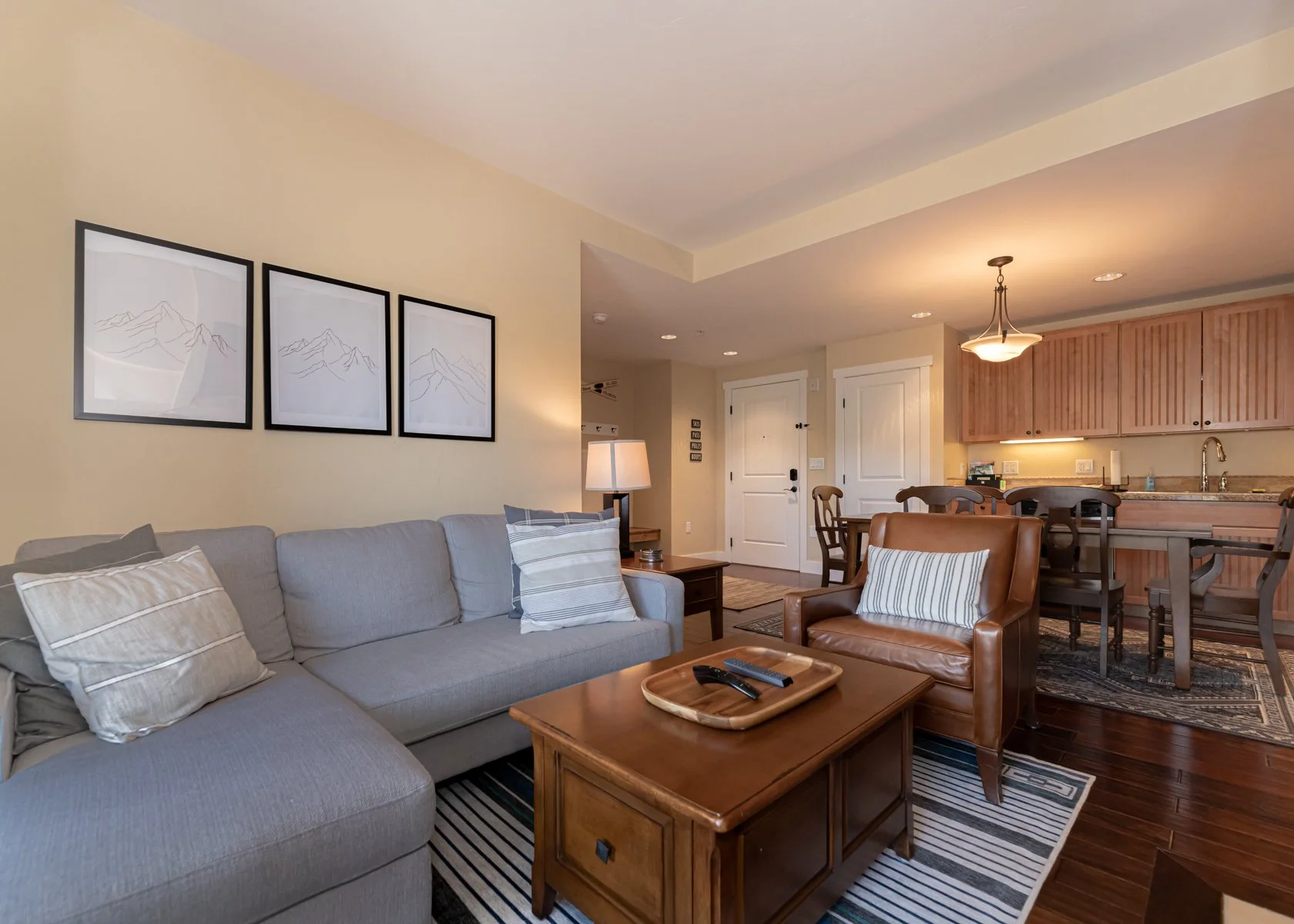 Living room with beige walls, gray couch with pillows, wooden coffee table with TV remote, leather armchair with pillow, and framed mountain artwork on the wall. Kitchen with wooden cabinets and dining table in background.