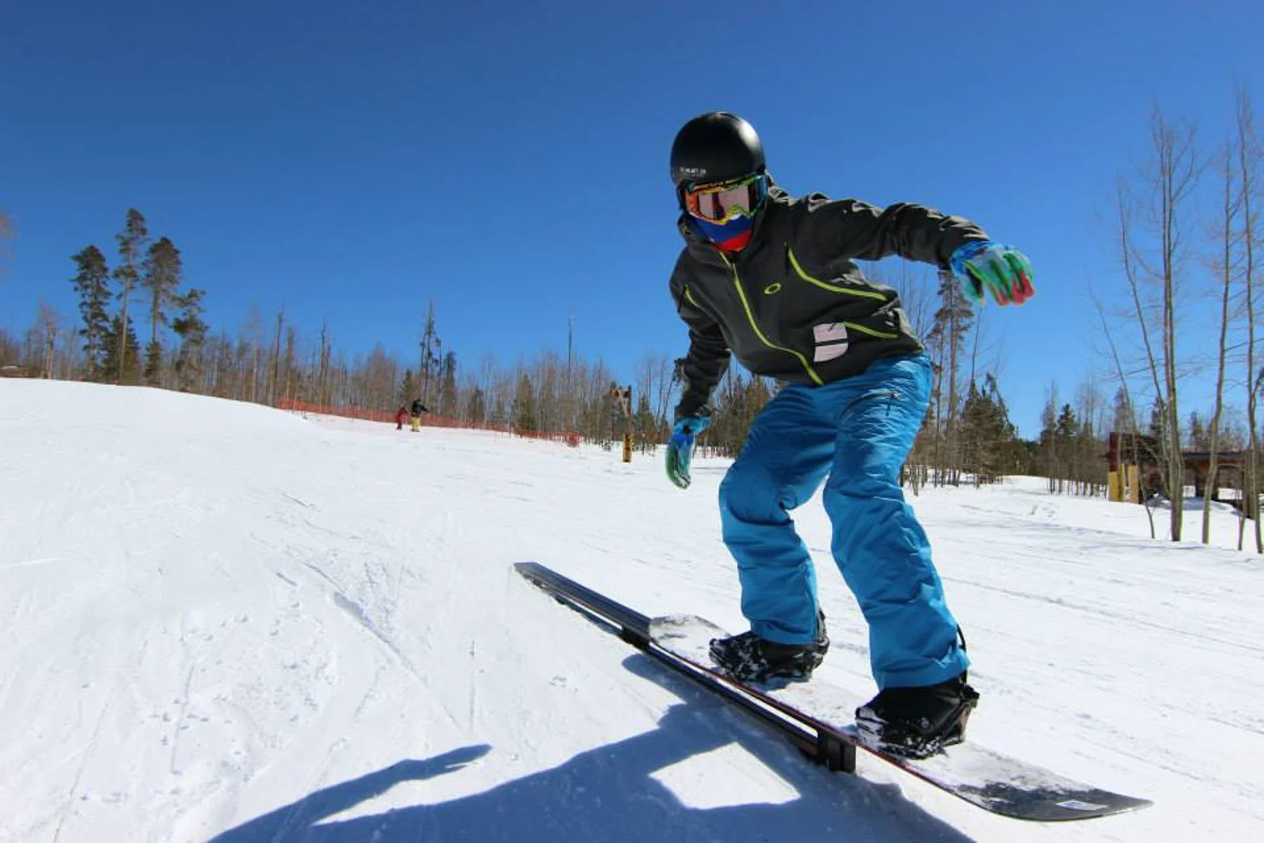 A person in snowboarding gear, including a black helmet, goggles, a black jacket with yellow accents, blue pants, and gloves, is snowboarding on a snowy slope under a clear blue sky with leafless trees in the background.
