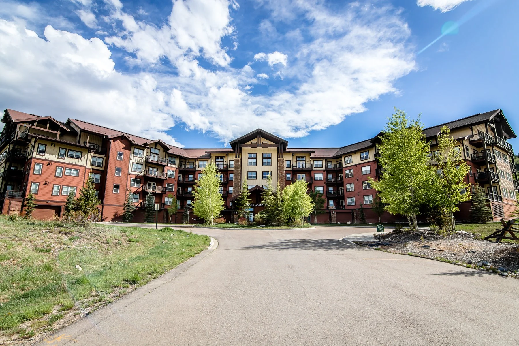 A multi-story residential building with a curved facade, balconies, and a parking garage at the base, surrounded by green trees and a paved driveway under a partly cloudy blue sky.
