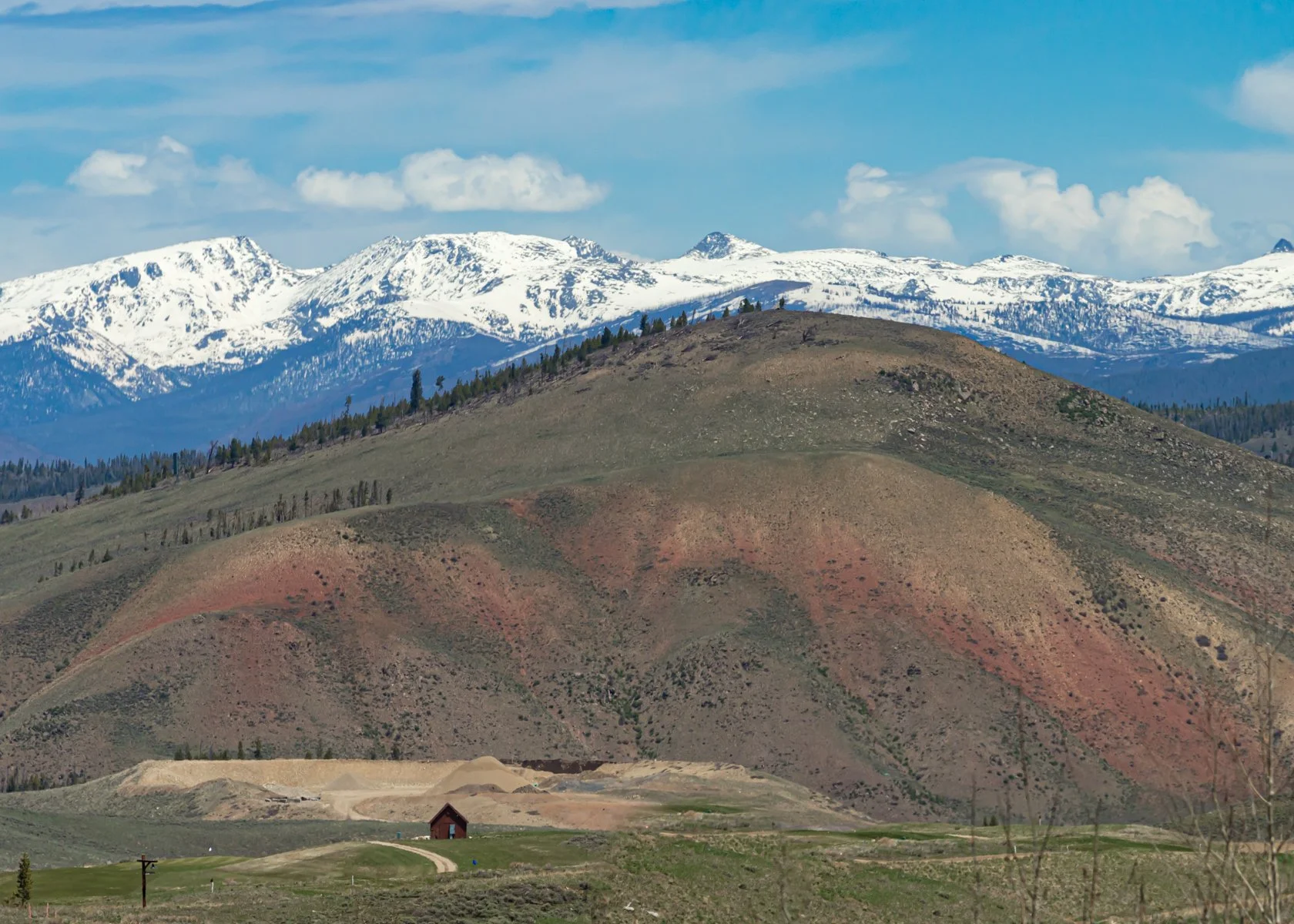Scenic landscape featuring rolling green hills in the foreground and snow-capped mountains in the background under a partly cloudy blue sky.