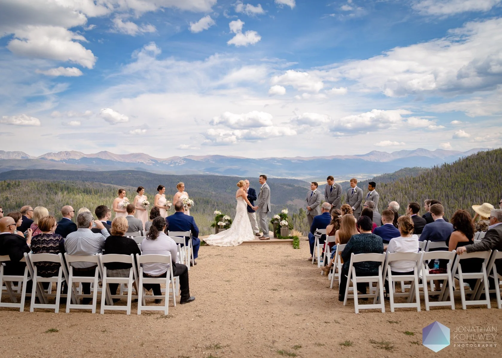 A wedding ceremony outdoors with a mountain view, bride and groom holding hands at altar, surrounded by bridesmaids and groomsmen, seated guests, clear sky and mountains in background.