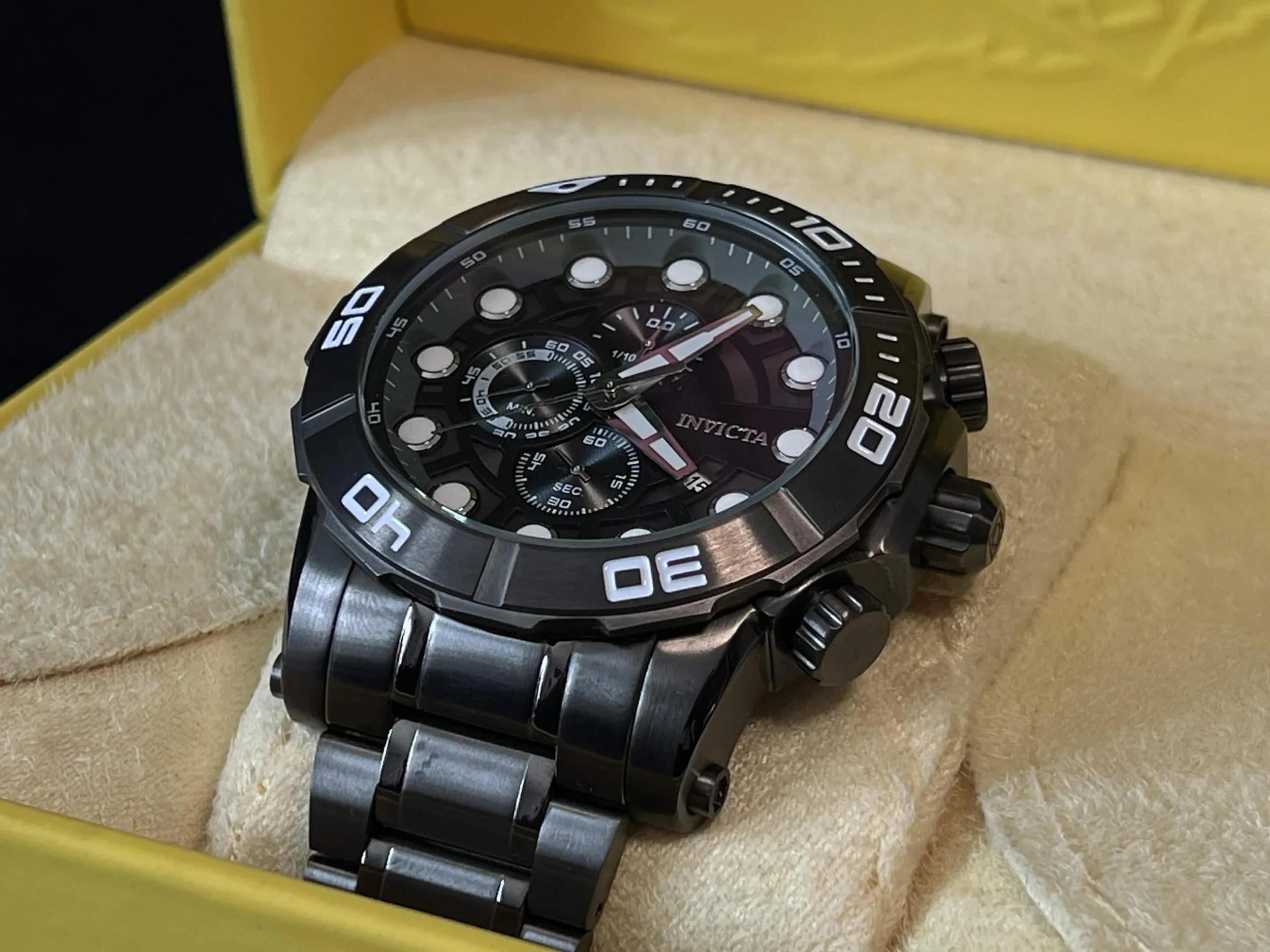 A black and silver wristwatch with a black bezel, large white markings, and multiple dials, placed on a yellow and beige textured surface inside a yellow box.