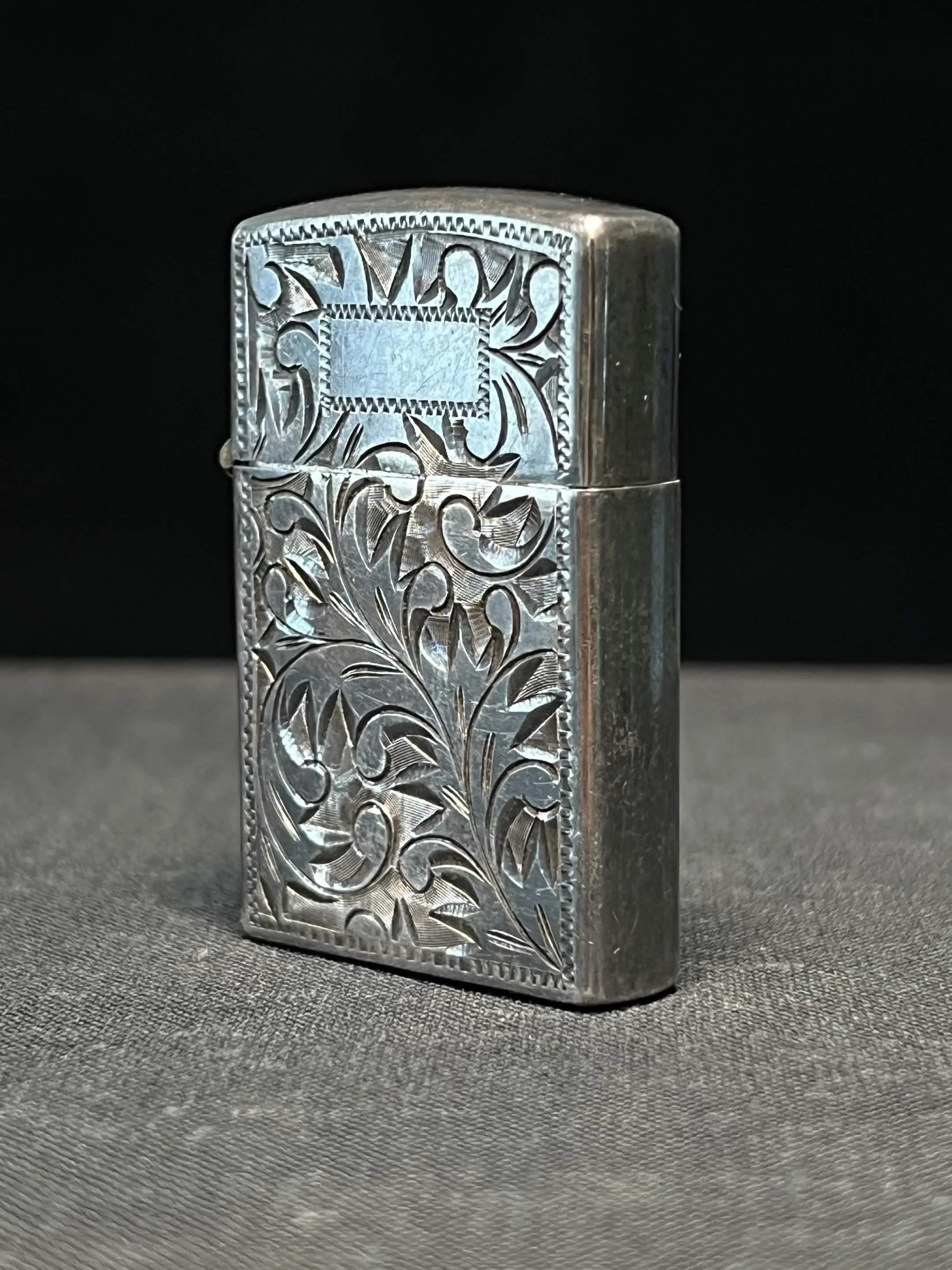 Close-up of a silver engraved cigarette lighter with floral and geometric patterns, placed on a textured dark surface.