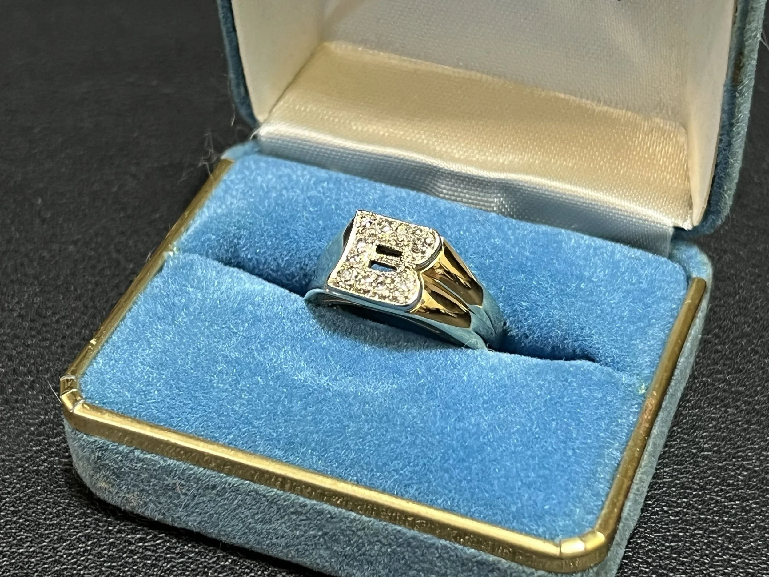 A gold ring with a large number of small diamonds on it, resting inside a white and blue jewelry box with a gold border, lined with blue velvet.