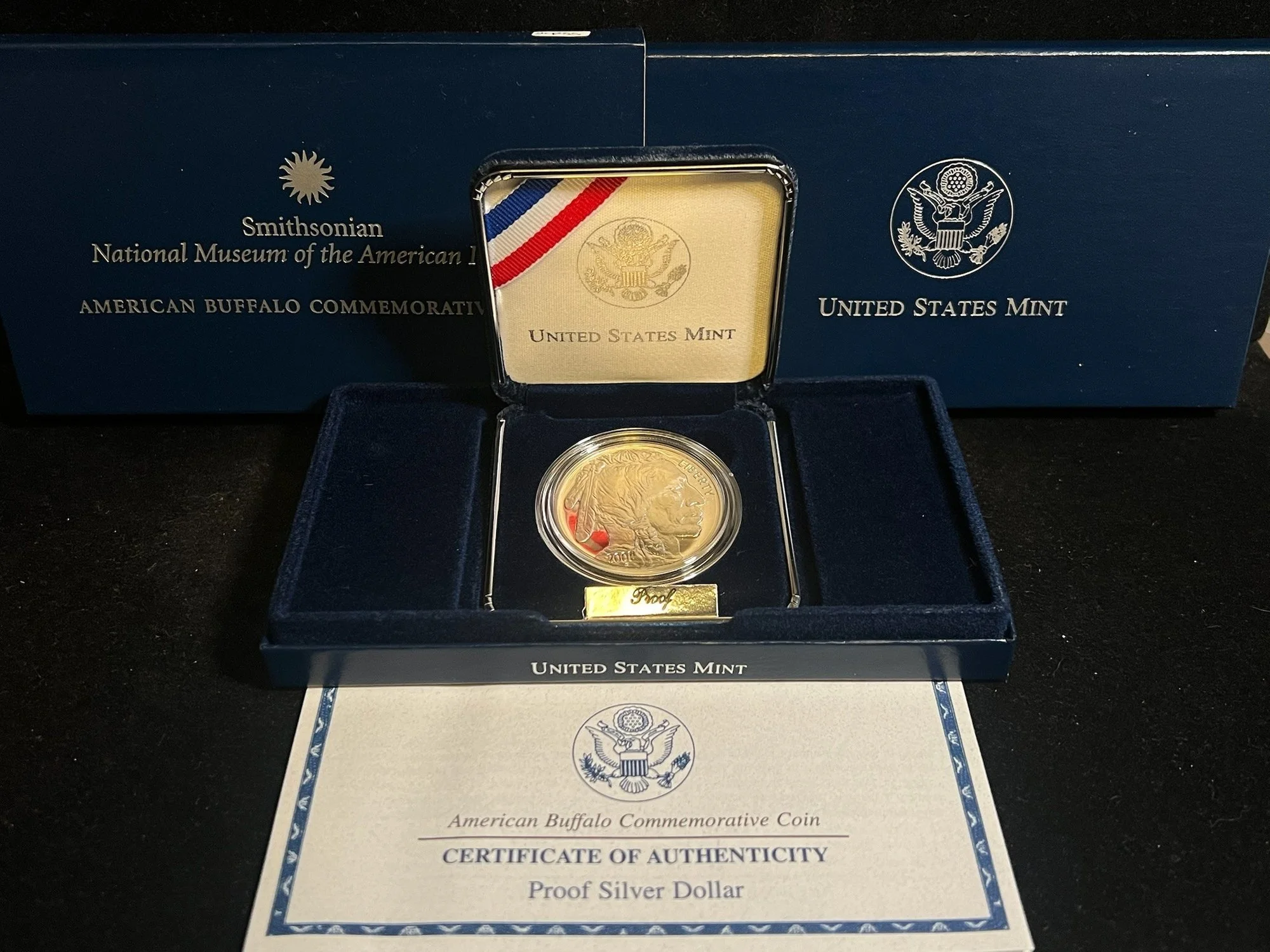 A commemorative silver dollar coin displayed in a case, with a certificate of authenticity from the United States Mint, celebrating the American Buffalo and related with the Smithsonian National Museum of the American Indian.