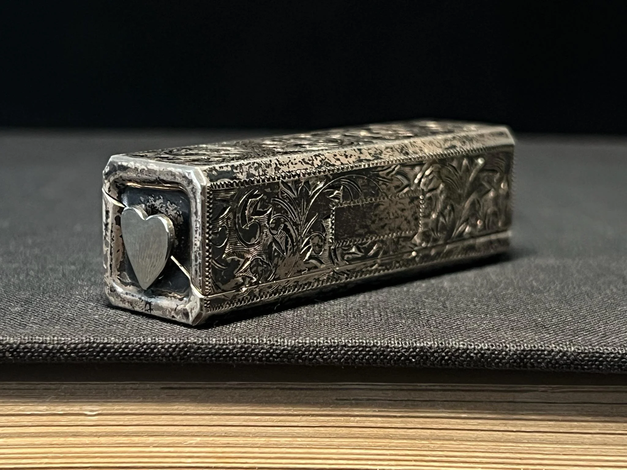 A silver engraved object with floral patterns and a heart shape on the front, resting on a dark textured surface with a wooden strip at the bottom.