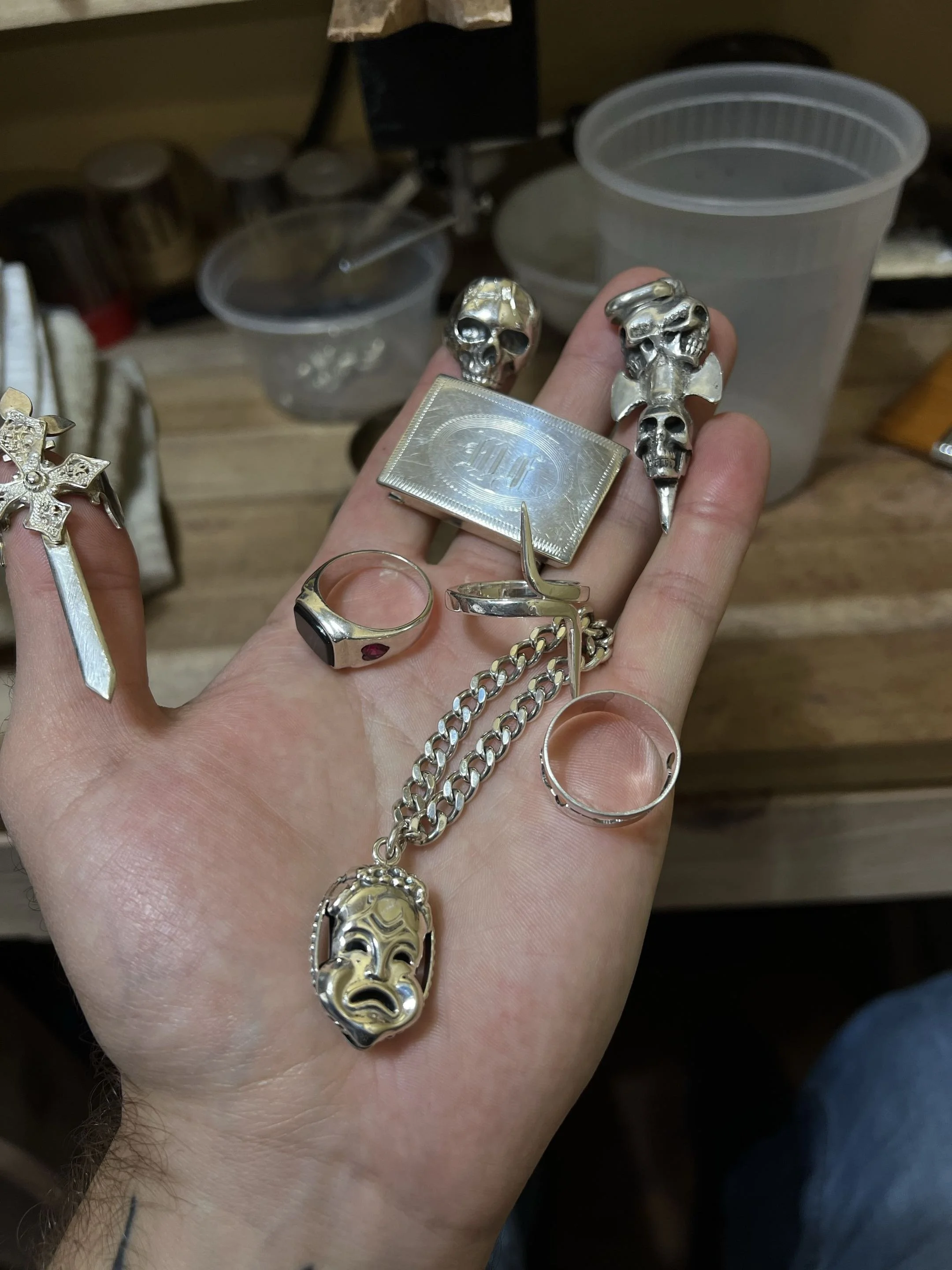 Close-up of a person's hand holding various silver jewelry pieces, including rings, earrings, and skull-shaped accessories, against a background of plastic containers and kitchen items.