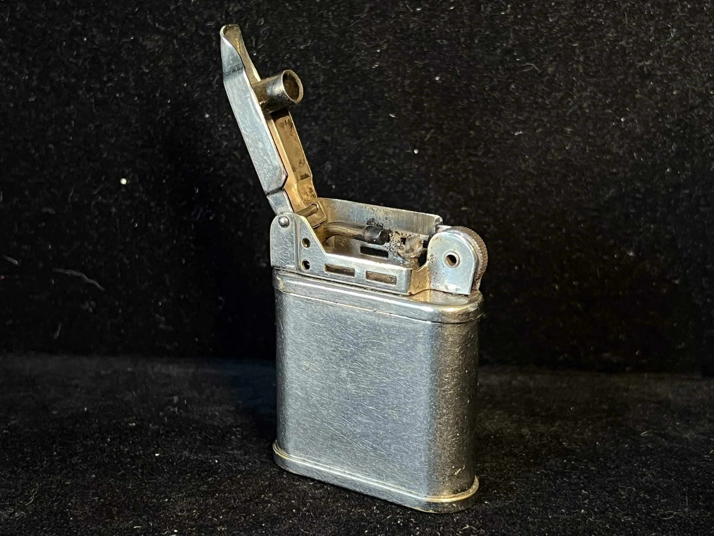 A metallic lighter with an open lid, placed on a black textured surface.