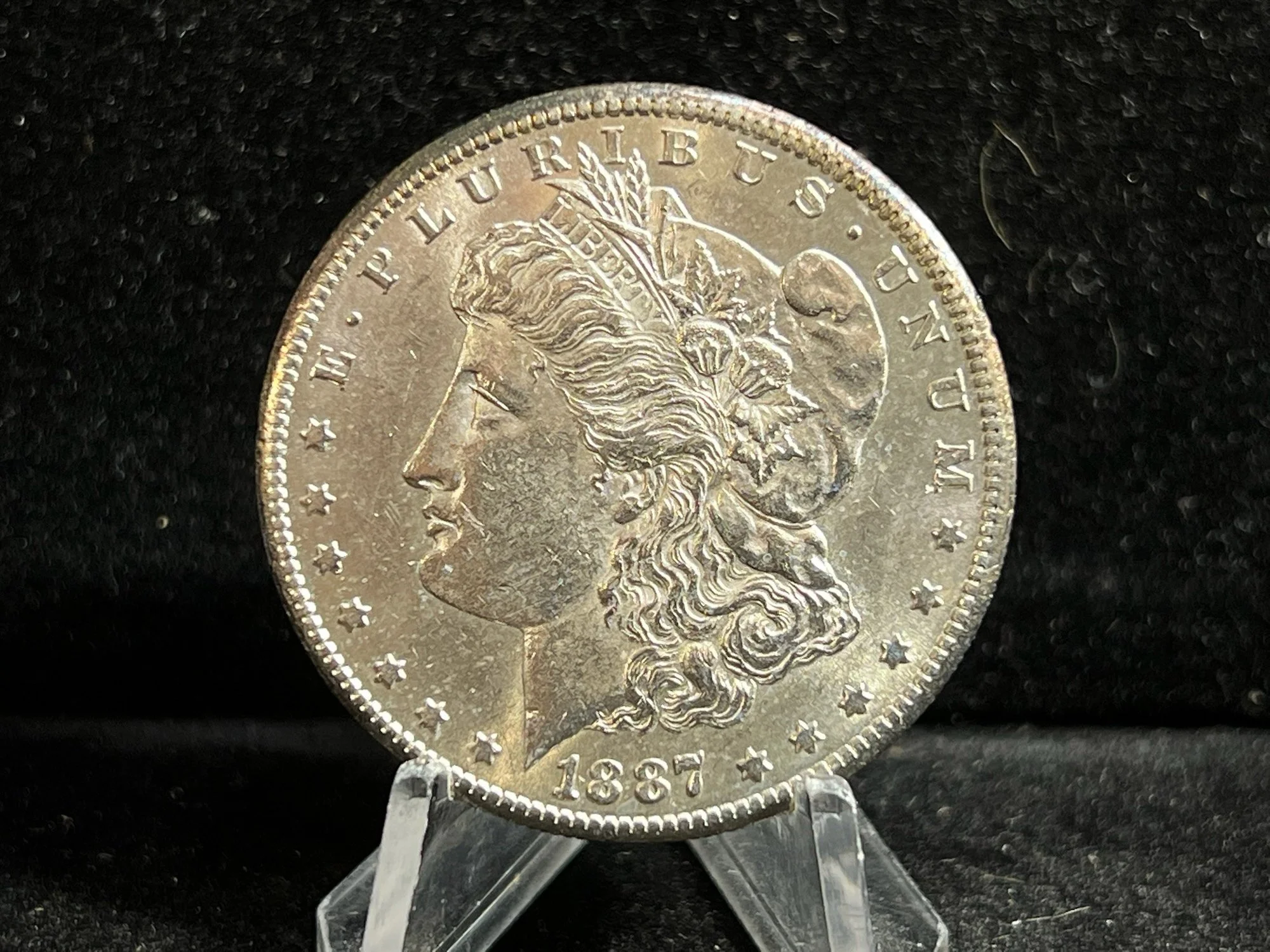 A 1887 Morgan silver dollar coin placed on a small metal stand against a black textured background.