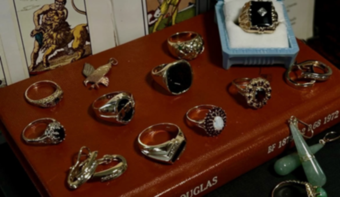 A collection of rings, a brooch, and a ring box displayed on a wooden surface, with a decorative box and artwork in the background.