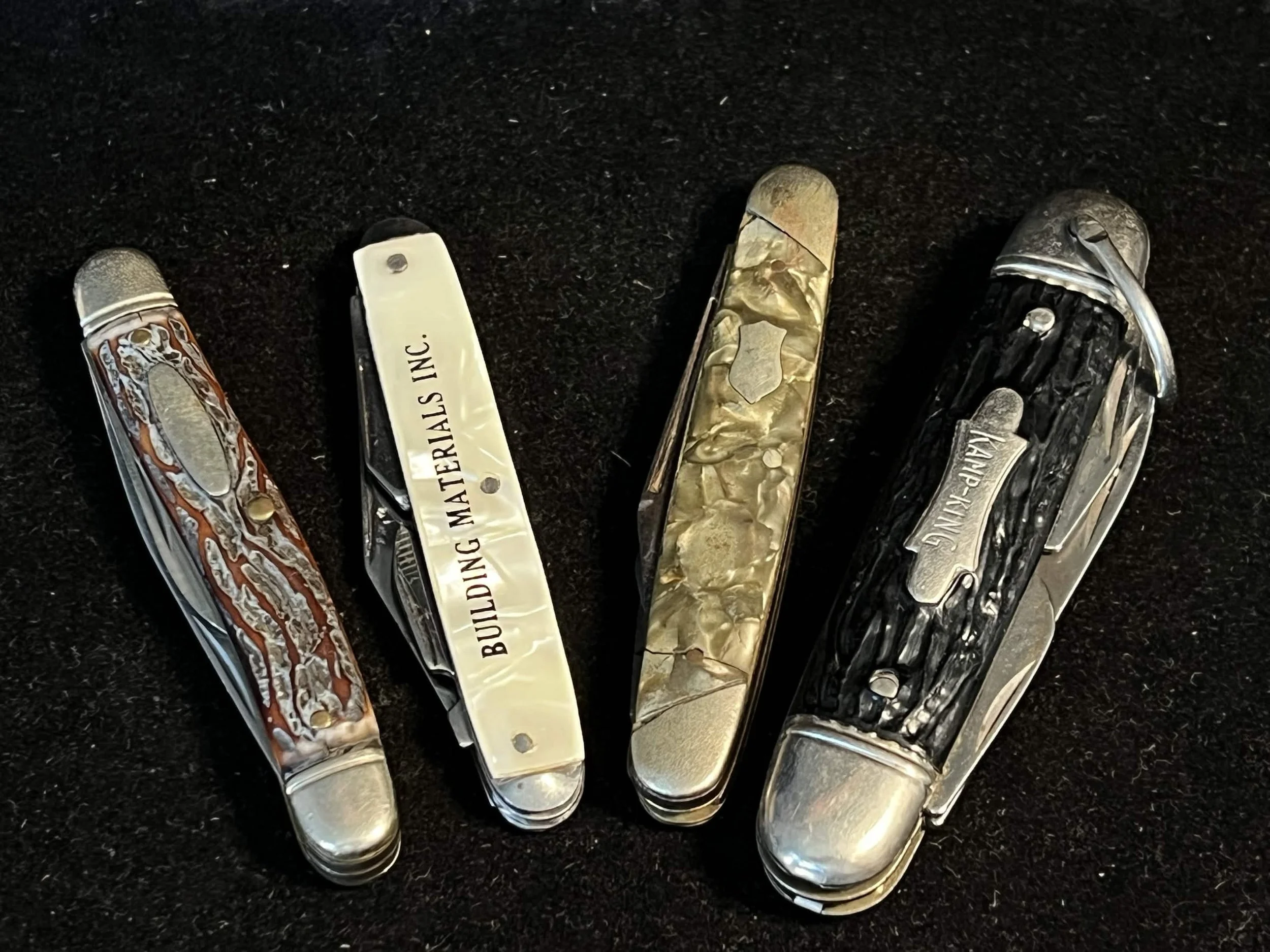 Collection of five pocket knives with various patterned and solid handles, some engraved with text, laid out on a black surface.