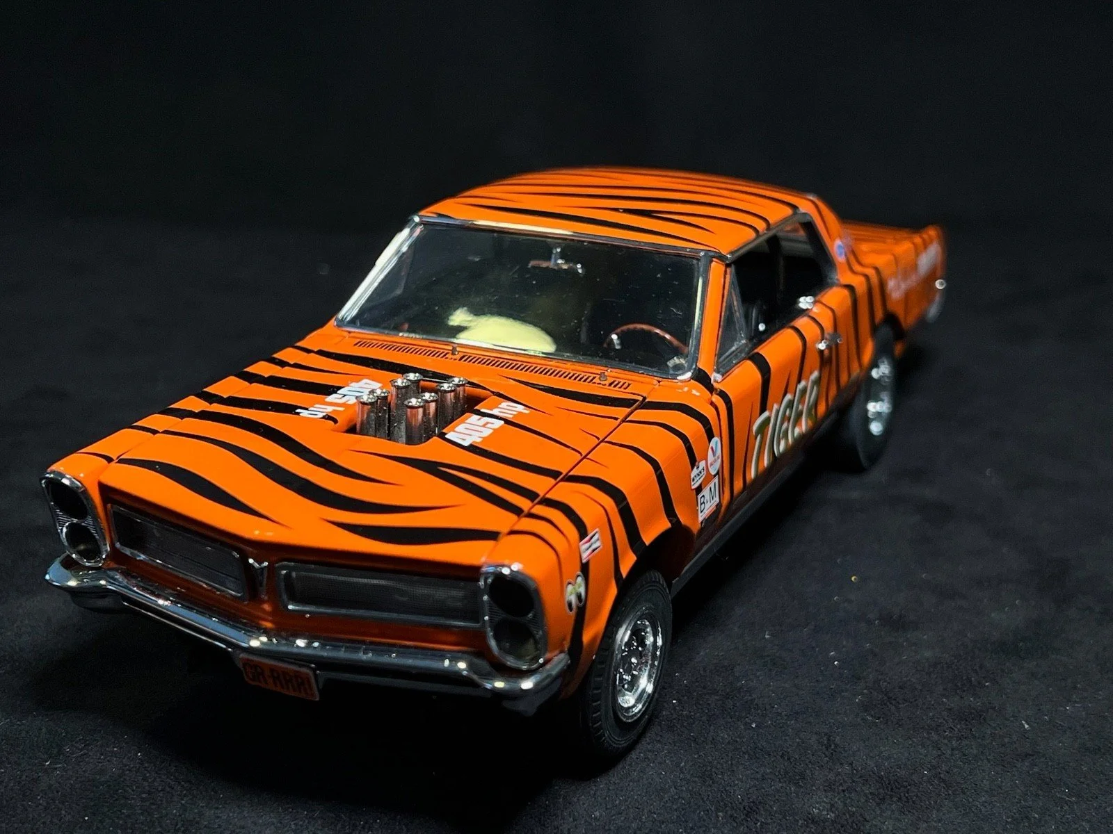 Close-up of a model car with a bright orange paint job and black tiger stripe pattern, featuring detailed engine parts on the hood and various stickers and decals.