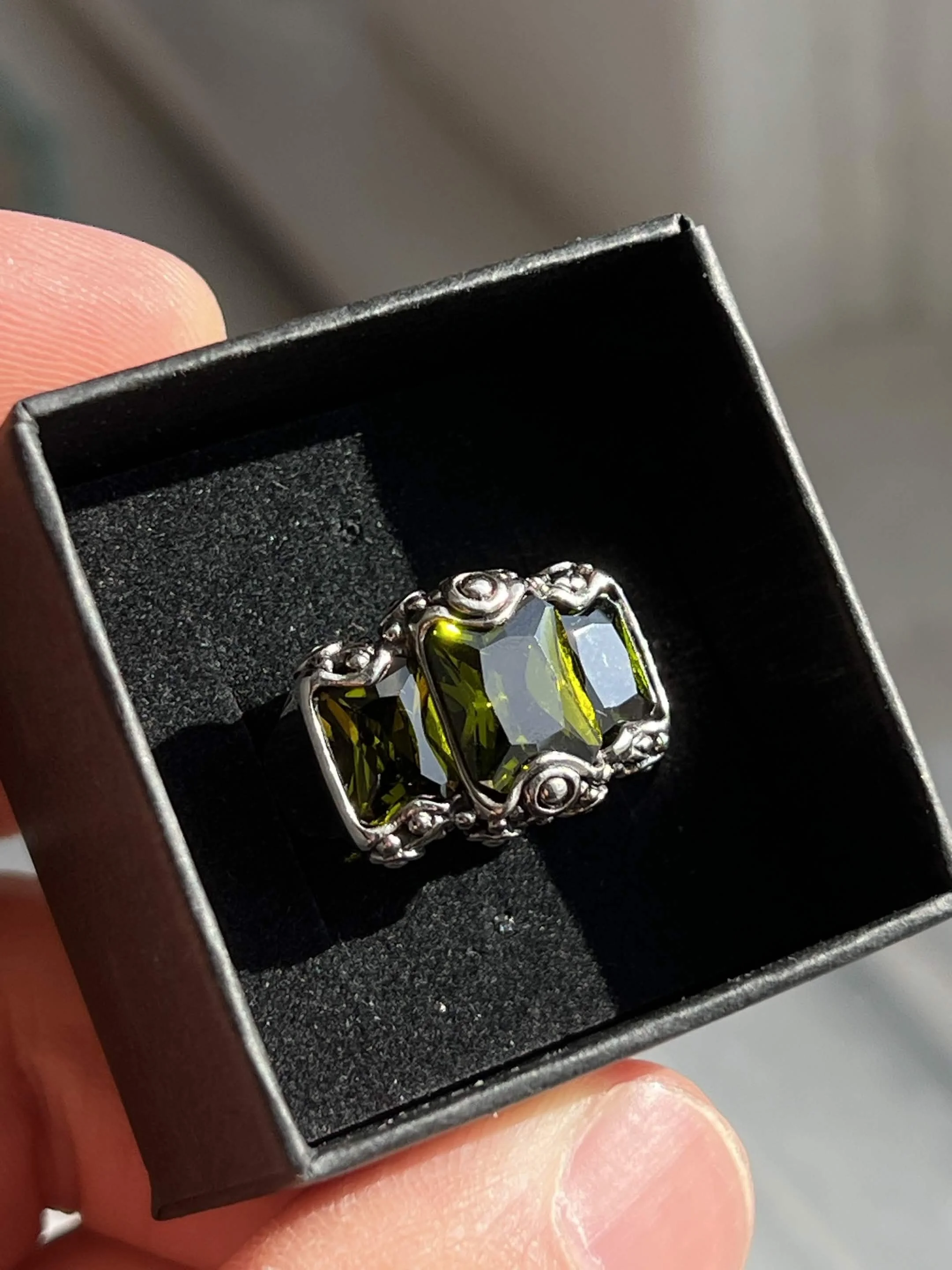 A person's hand holding a small jewelry box containing a ring with a large, rectangular, faceted green gemstone and a decorative silver band.