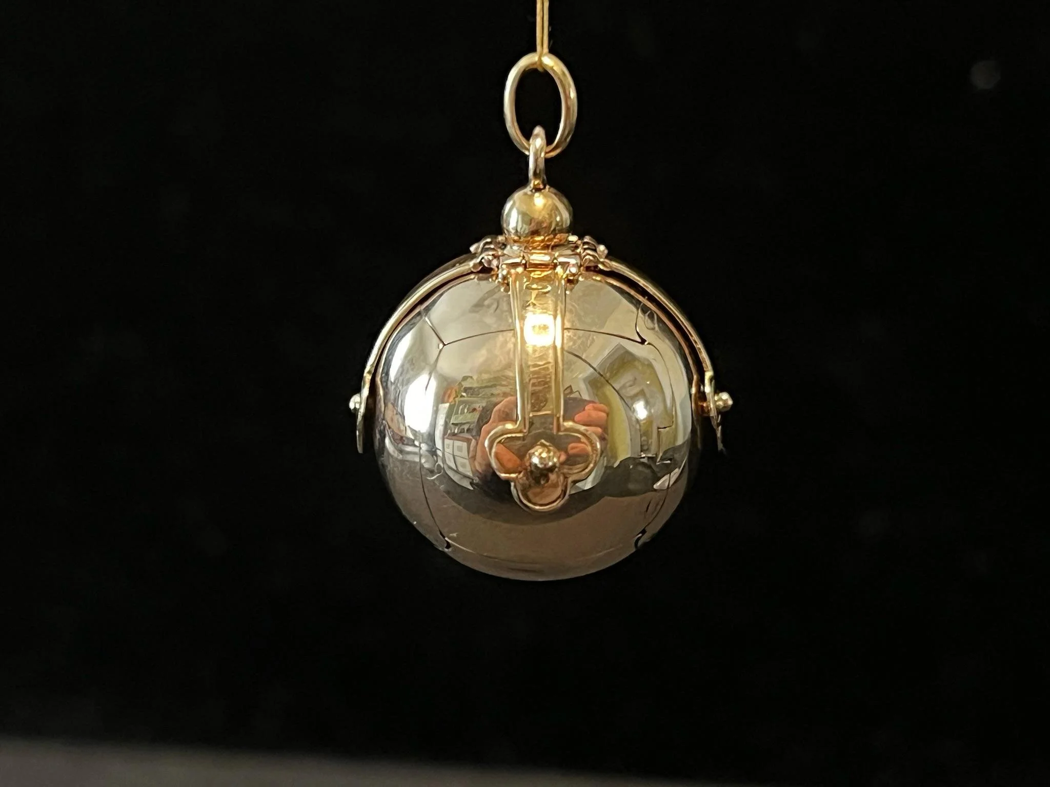 A teardrop-shaped pendant with a reflective silver surface, gold accents, and a small loop at the top for hanging, set against a black background.