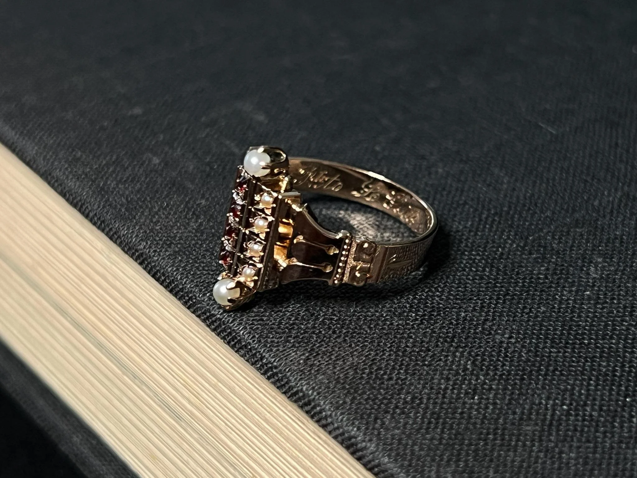 Close-up of a decorative ring with a pearl and red gemstones, resting on a black textured surface next to a light-colored wooden surface.
