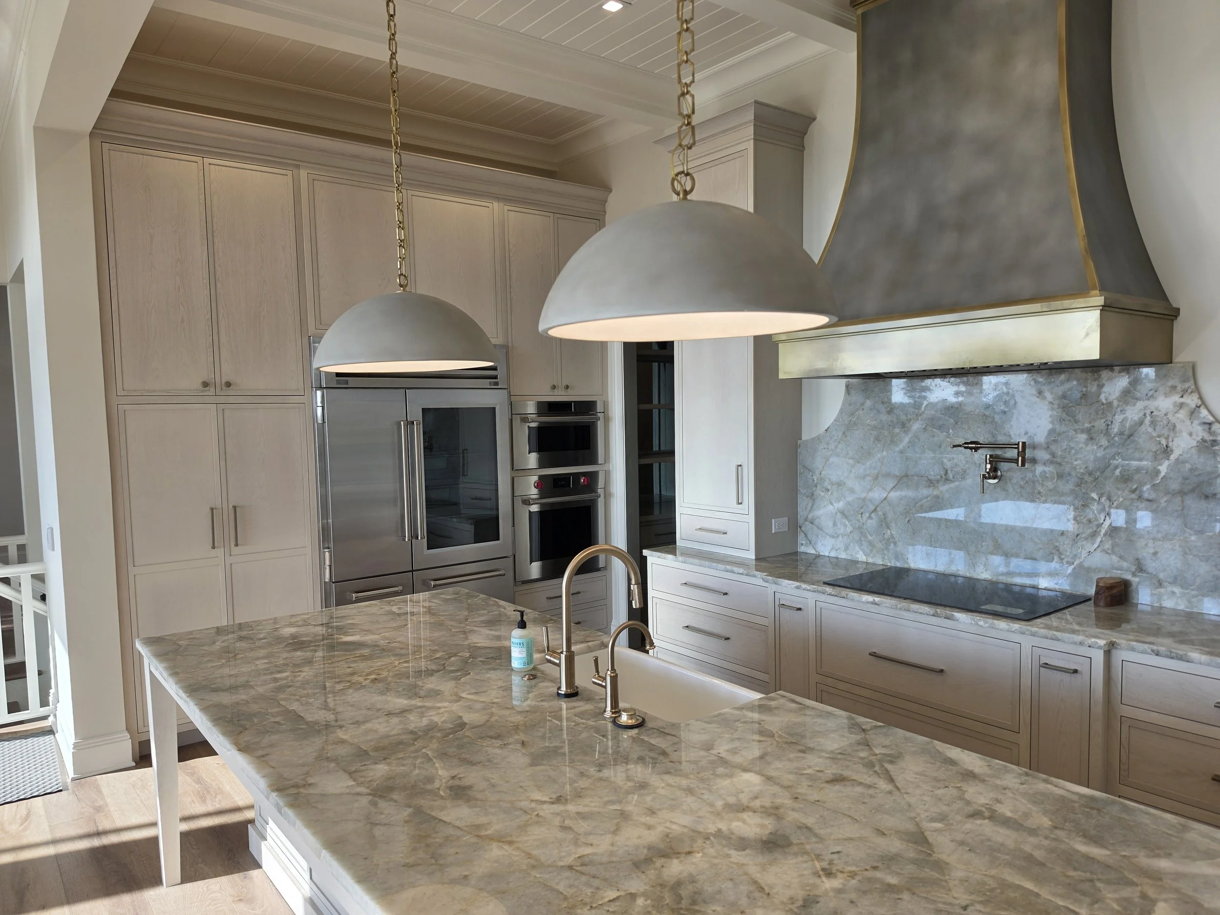 A spacious kitchen with a large marble island, beige cabinets, stainless steel refrigerator, oven, and microwave. Two large pendant lights hang above the island. The kitchen has a marble backsplash and a modern faucet at the sink.