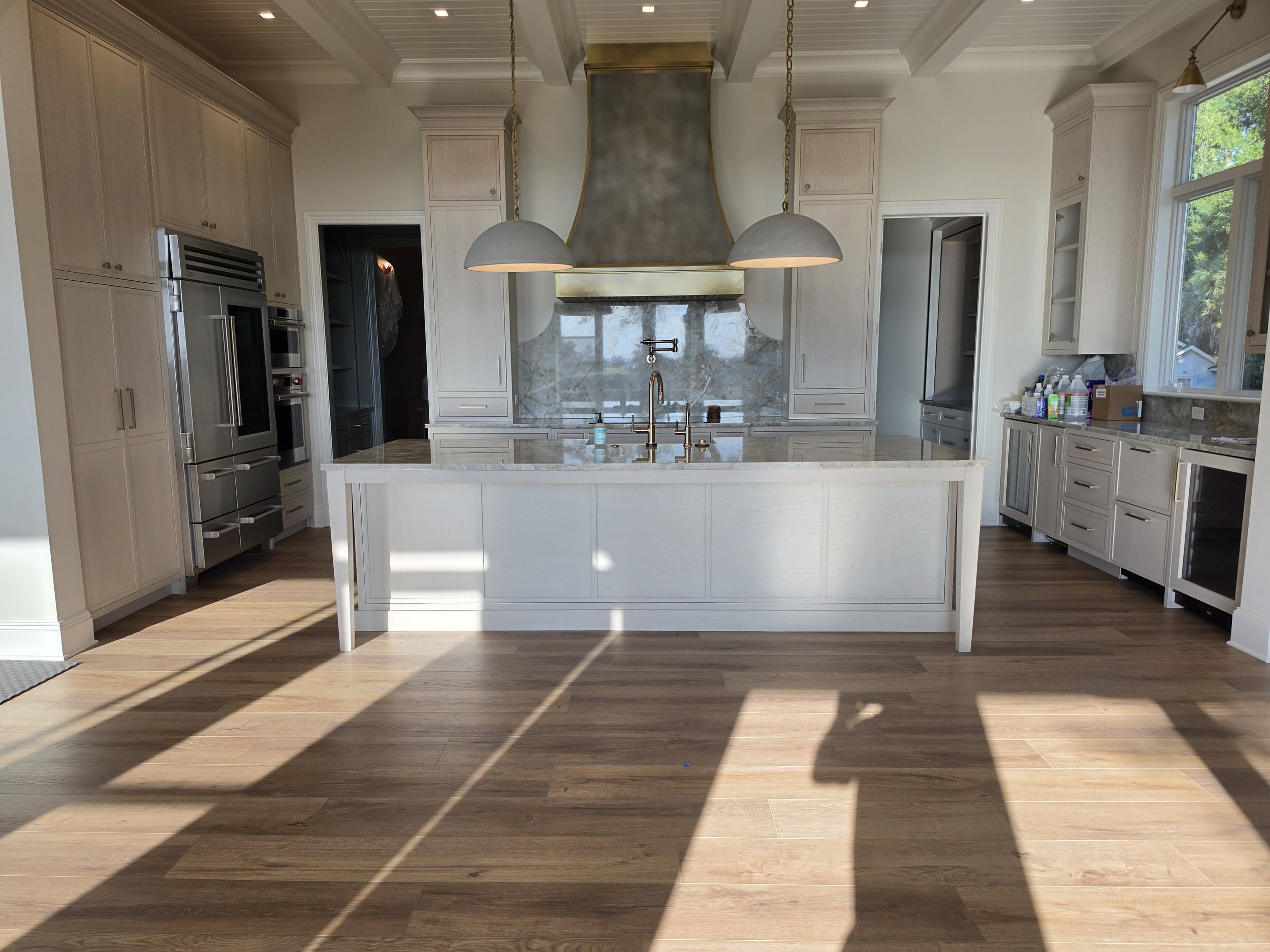 A spacious kitchen with white cabinets, a marble backsplash, a kitchen island with a sink, two hanging pendant lights, stainless steel appliances, and hardwood floors, illuminated by natural light from large windows.