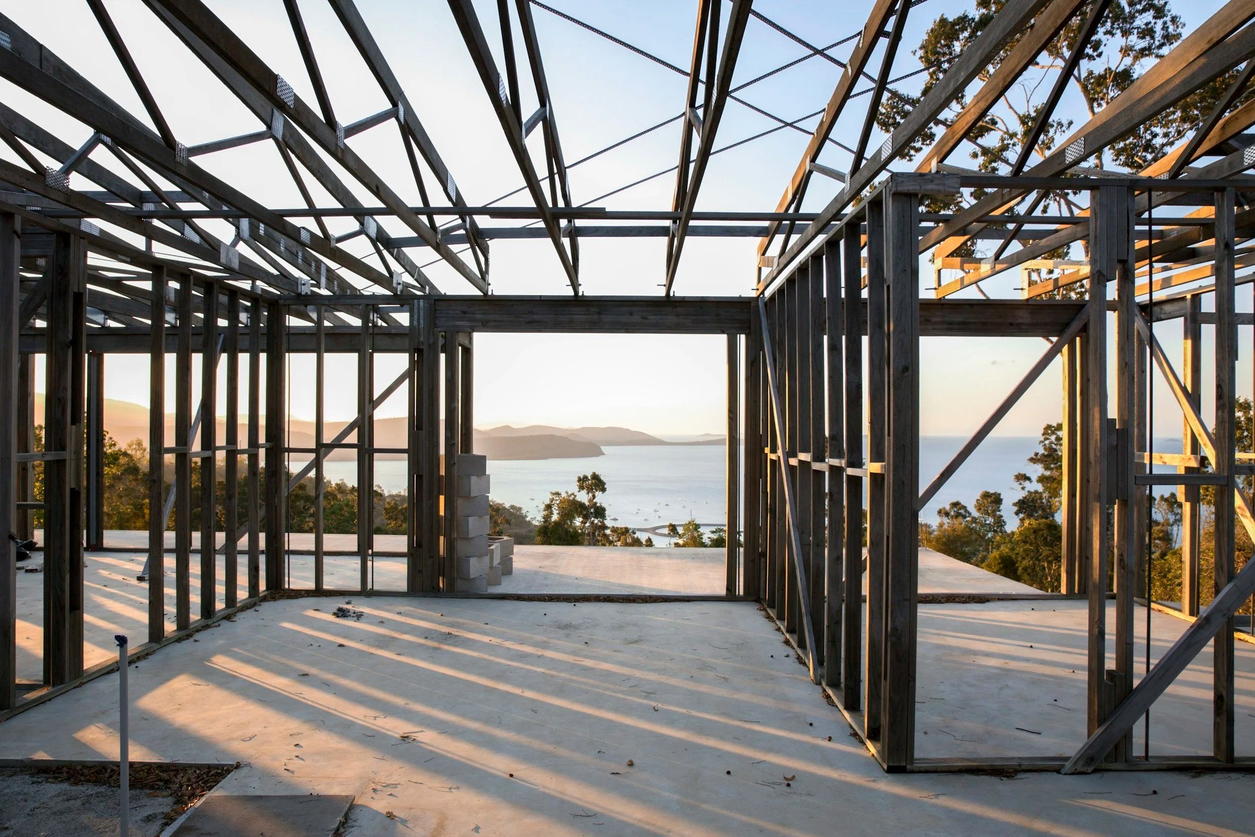 The image shows the framework of a house under construction with wooden beams, with a scenic view of the ocean and distant hills in the background, during sunset.