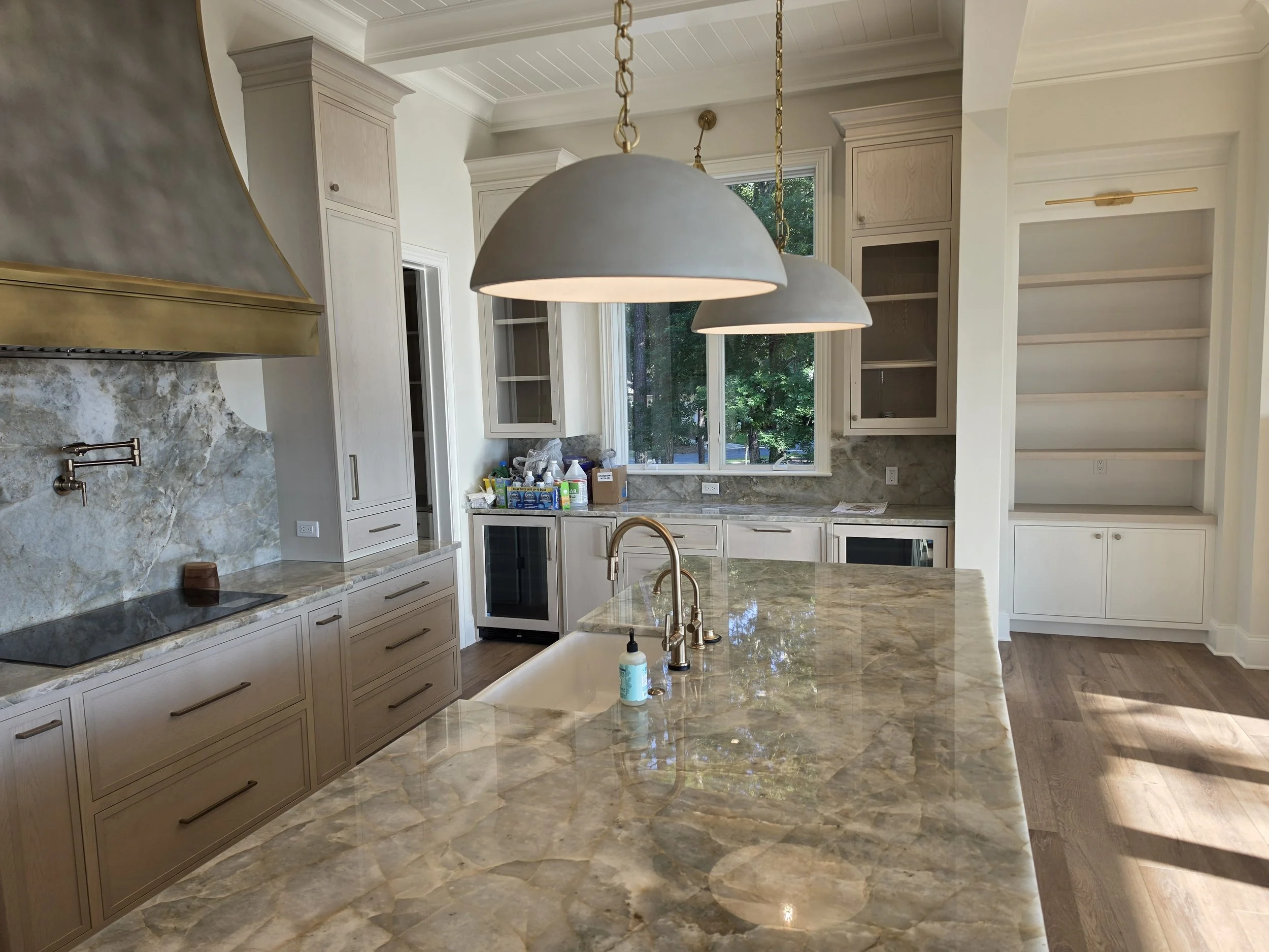 Interior view of a modern kitchen with beige cabinetry, a marble kitchen island, and large windows revealing greenery outside. Two large pendant lights hang over the island, and the kitchen appears to be in the process of being organized or renovated.