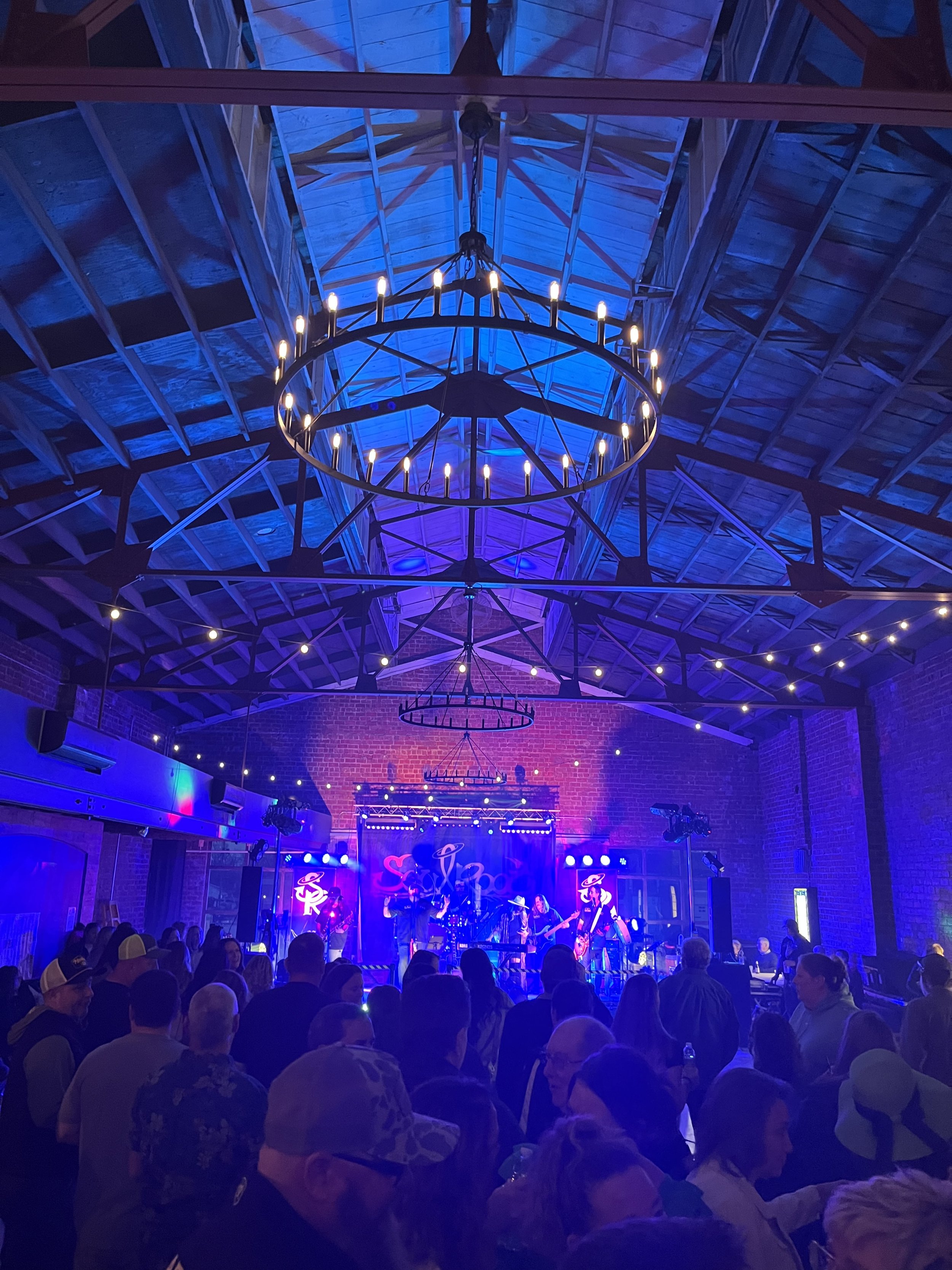 Indoor concert venue with a stage, musicians, and a crowd of people, illuminated by blue and purple lighting, featuring a high ceiling with exposed wooden beams and a large chandelier.