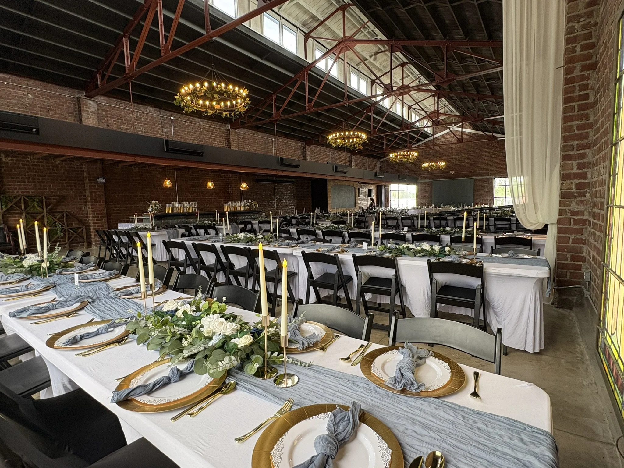 Long dining tables decorated with greenery, white roses, candles, and gold chargers in a rustic industrial venue with brick walls, high ceilings, large windows, and chandeliers.