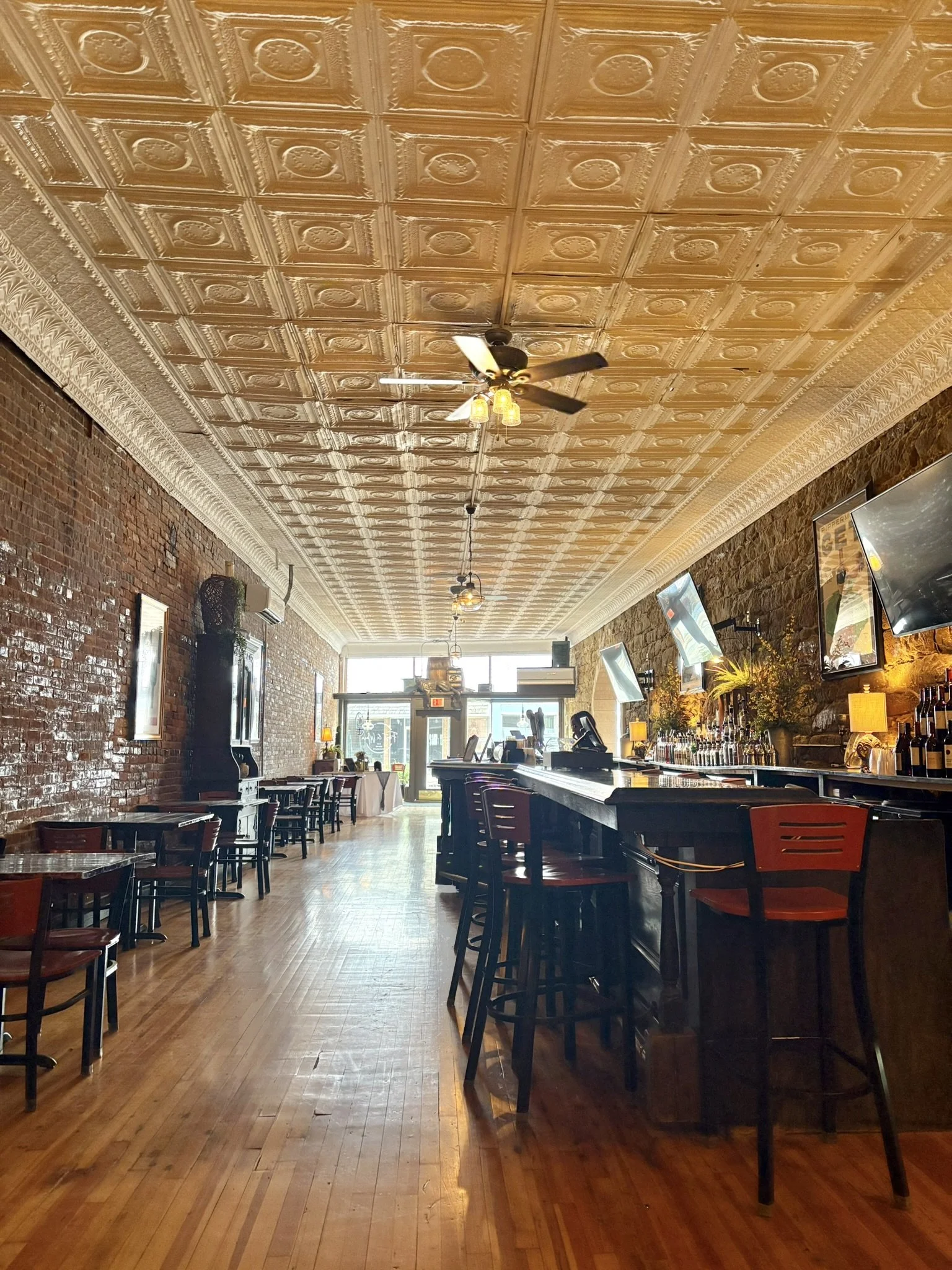 Interior of a restaurant or cafe with wooden floors, brick walls, decorative tin ceiling, ceiling fan, and multiple wall-mounted televisions. There are tables and chairs arranged along the space, with a bar counter on the right side adorned with bott
