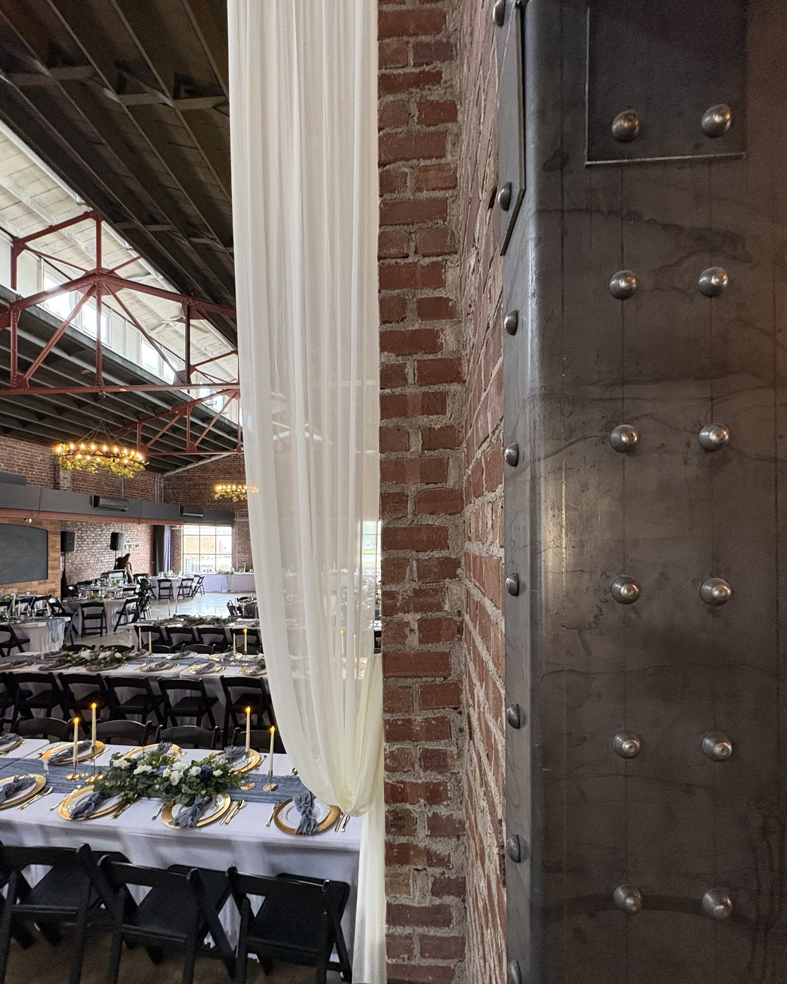 Indoor banquet hall decorated for an event with round tables, candles, elegant tableware, and floral centerpieces, featuring exposed brick walls and industrial-style decor.