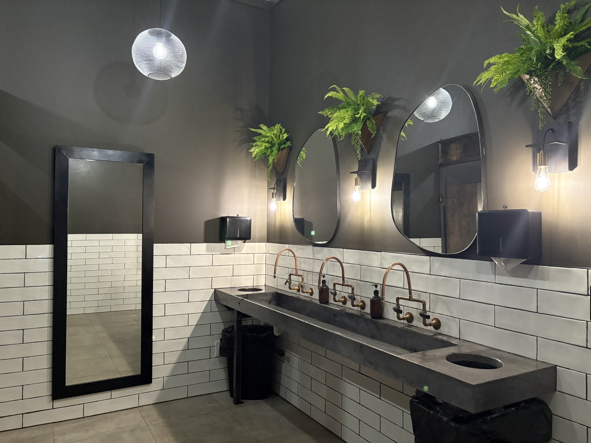 Modern bathroom with a large mirror, black fixtures, white subway tile walls, green plants, and industrial-style lighting.