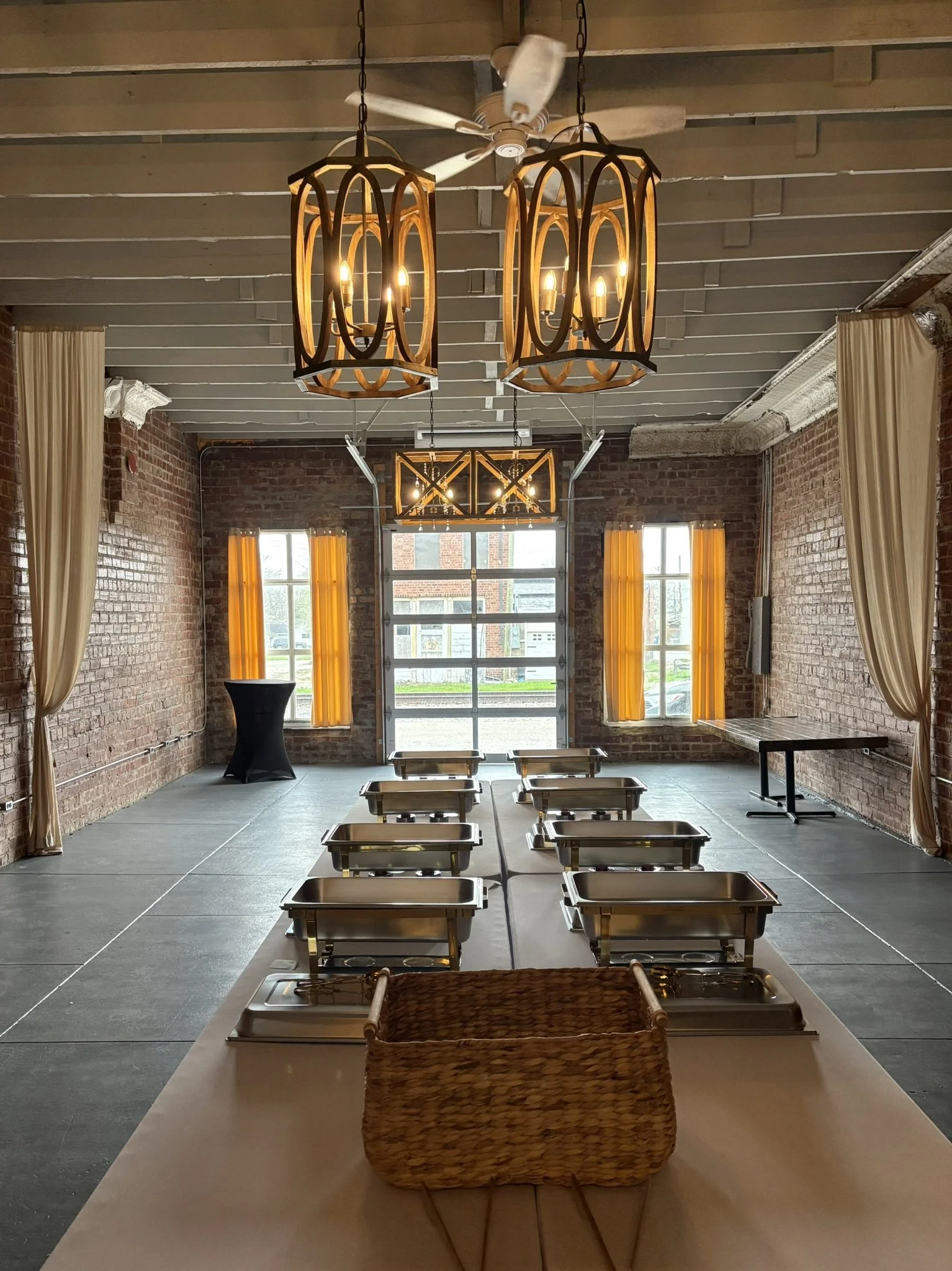 Interior of a rustic event space with brick walls, windows with yellow curtains, hanging chandeliers, and a ceiling fan. A table in the foreground has a wicker basket and a row of chafing dishes behind it. There is a black cocktail table to the left 