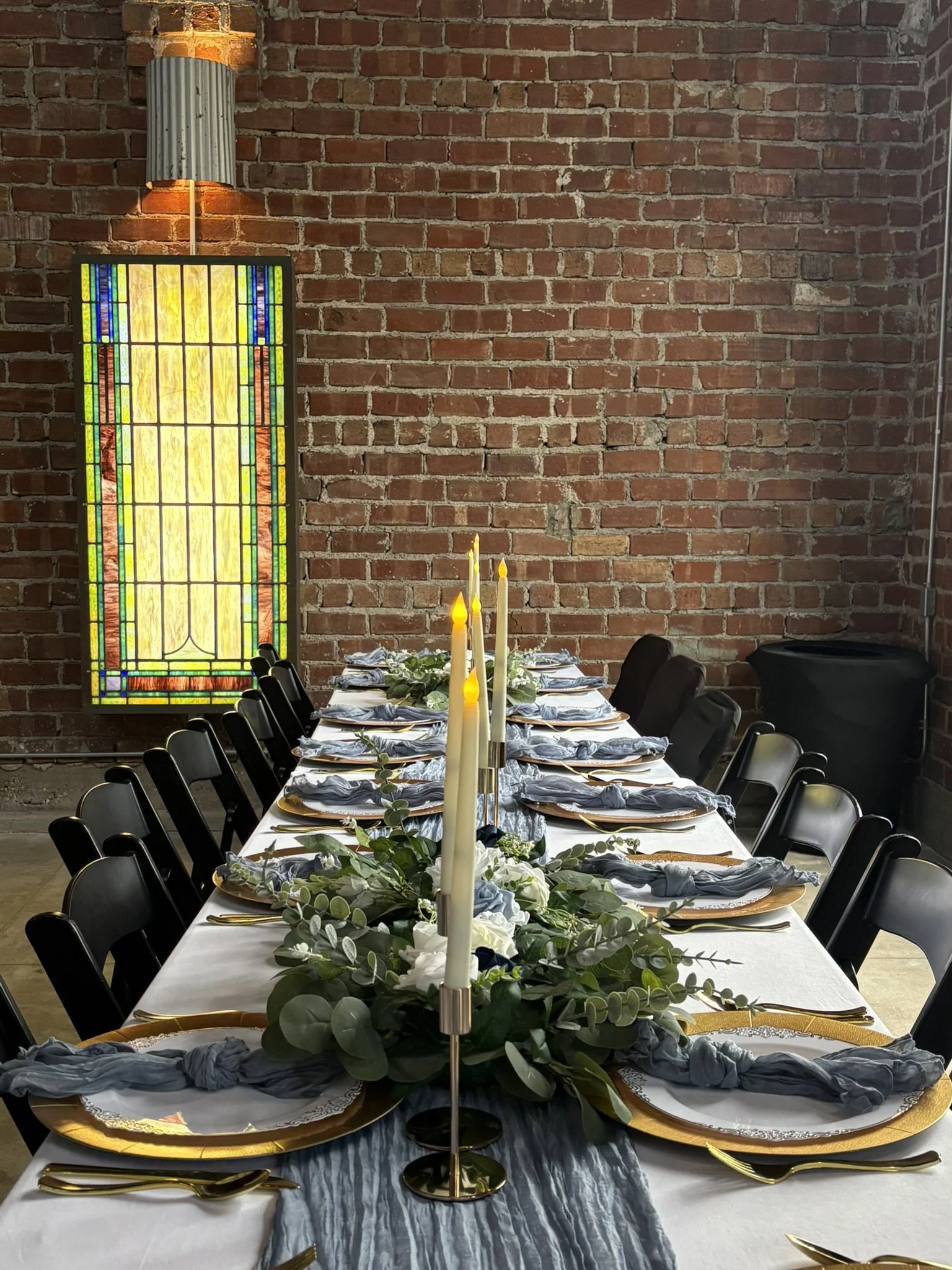 A long banquet table decorated for a special occasion with a white tablecloth, gold and black plates, gold utensils, and blue napkins. The center of the table features a floral greenery arrangement and tall white candles in gold holders. A stained gl