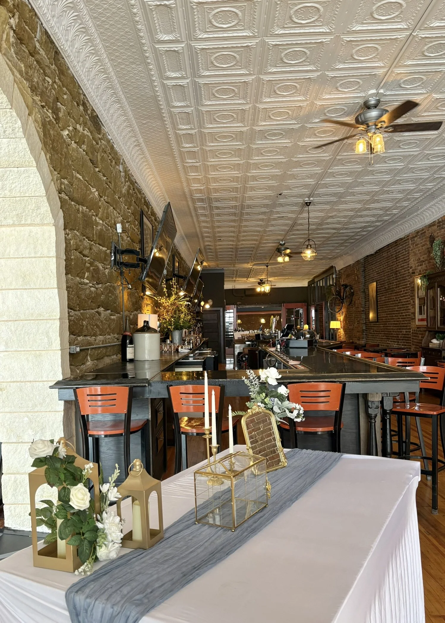 Interior of a restaurant or event space with a long table decorated with candles, lanterns, and floral arrangements, featuring brick walls, a patterned white ceiling, hanging lights, and a bar area in the background.