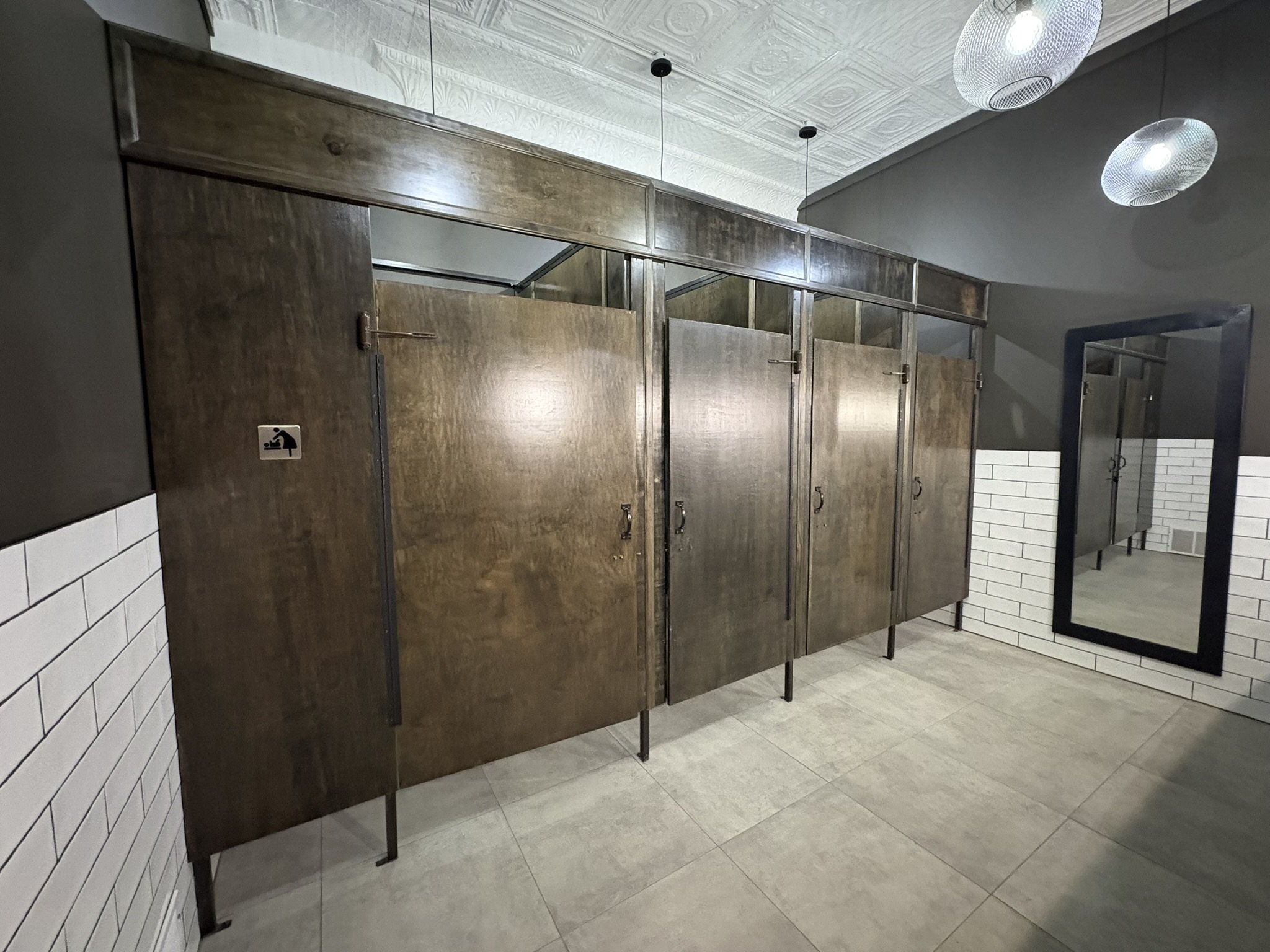 Public restroom with six wooden stall doors and a wall-mounted mirror. The stalls have metal handles, some with locks. The ceiling has decorative stamped tin pattern with two oval light fixtures. The floor is tiled in light gray, with a section of wh