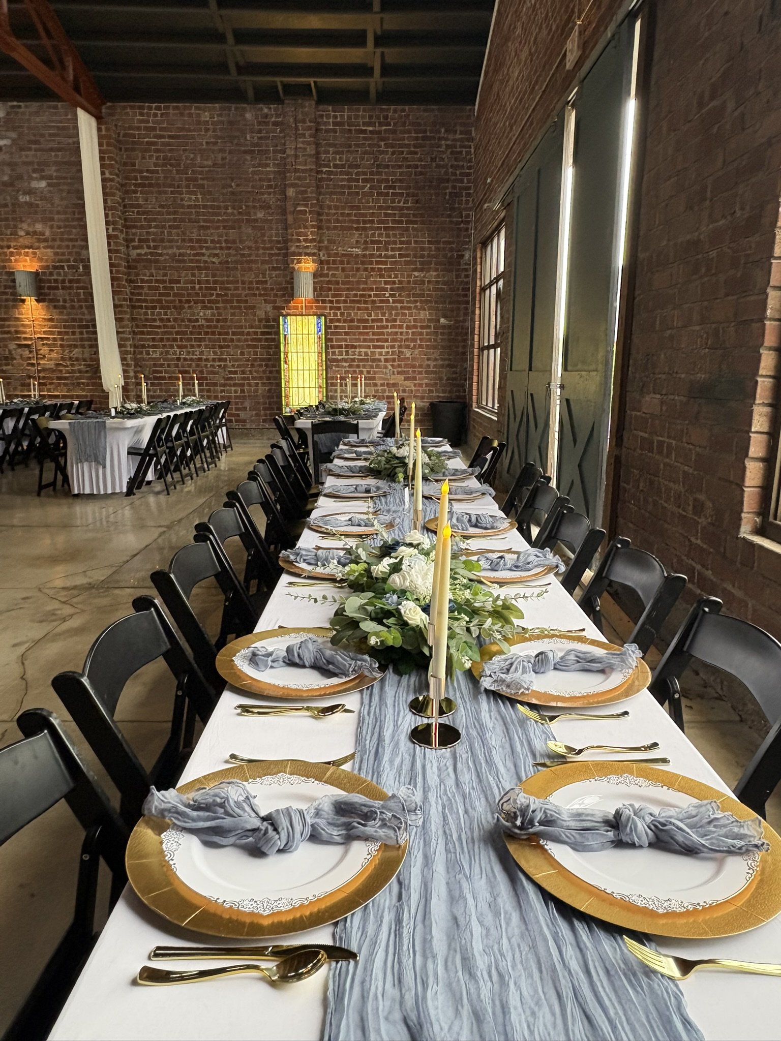 A long dining table in a rustic brick-walled room decorated for a formal event with gold and white plates, gray napkins, gold utensils, floral centerpieces, and yellow candles.