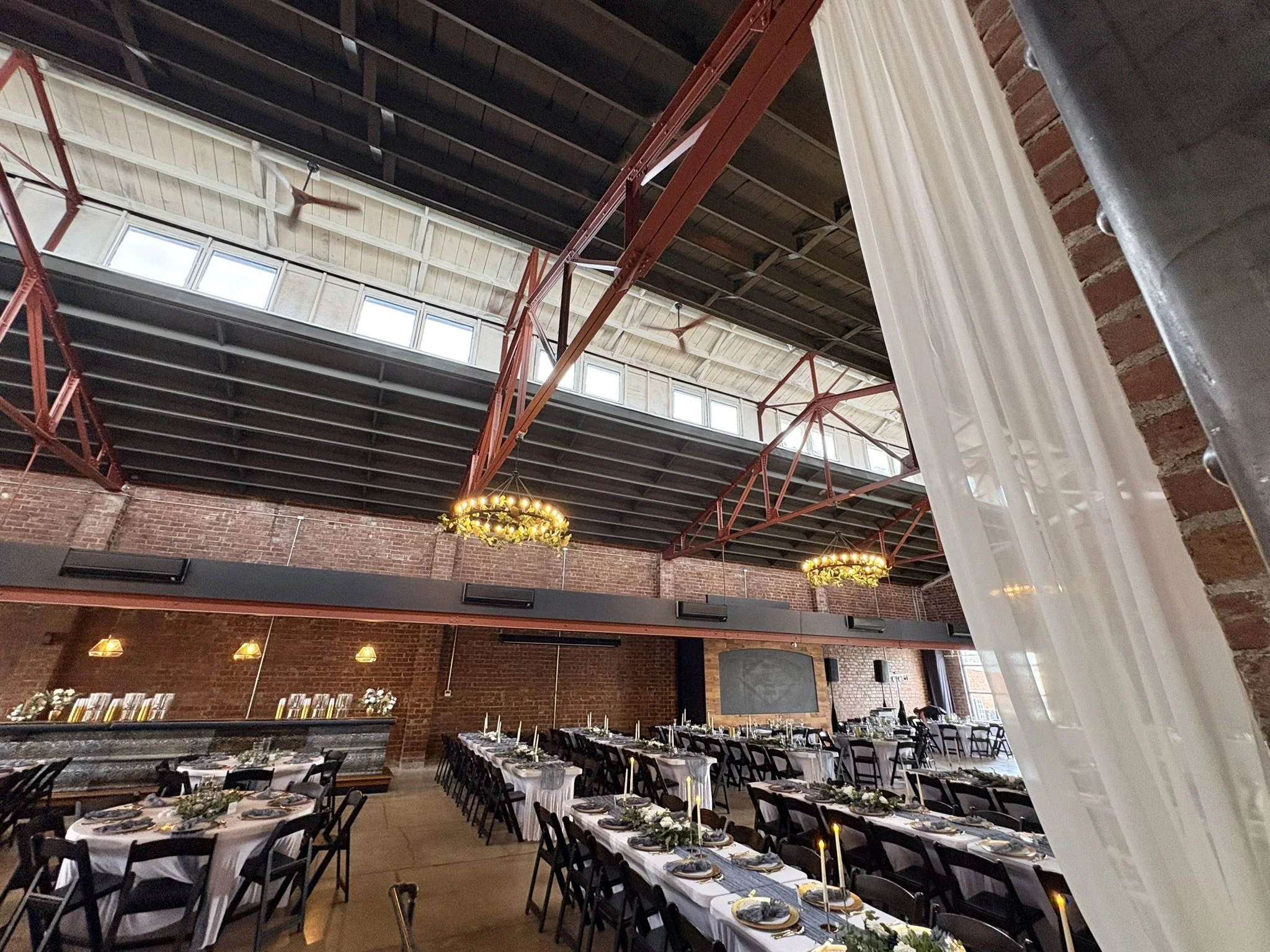 Long rectangular dining tables set with black chairs, white tablecloths, and candles, arranged in rows inside an industrial-style event space with brick walls, large windows, and chandeliers with greenery accents.