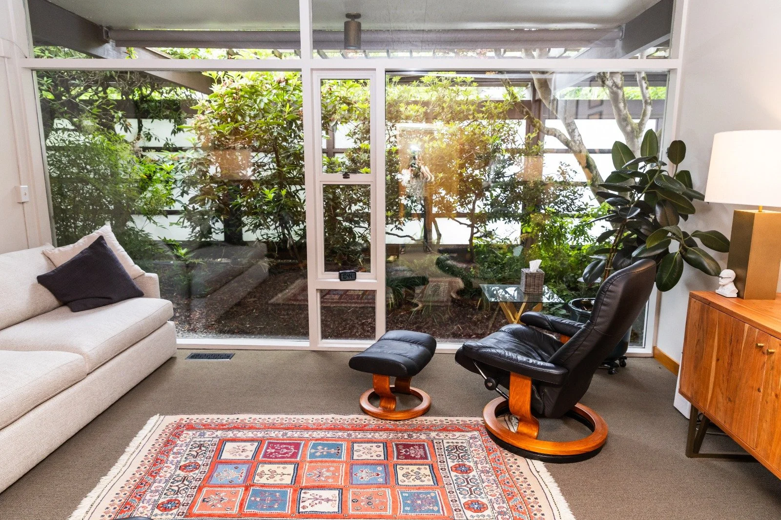 Living room with beige sofa, black leather chair with matching footstool, wooden cabinet, large indoor plant, and a glass wall revealing a lush outdoor garden with trees and bushes.