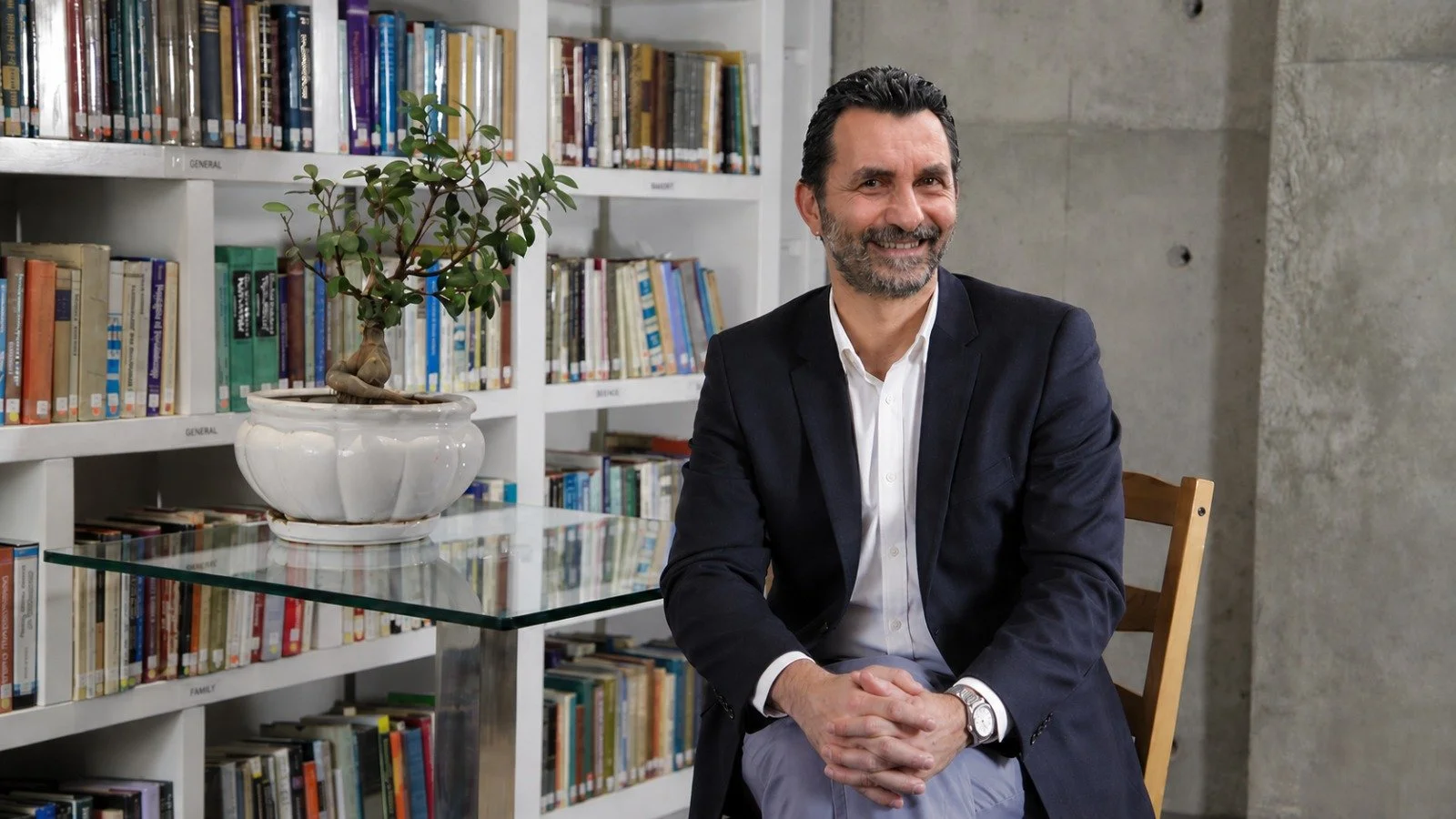 A man in a dark blazer and white shirt sitting on a wooden chair in a library, smiling at the camera, with a bookshelf and a potted plant on a glass table behind him.
